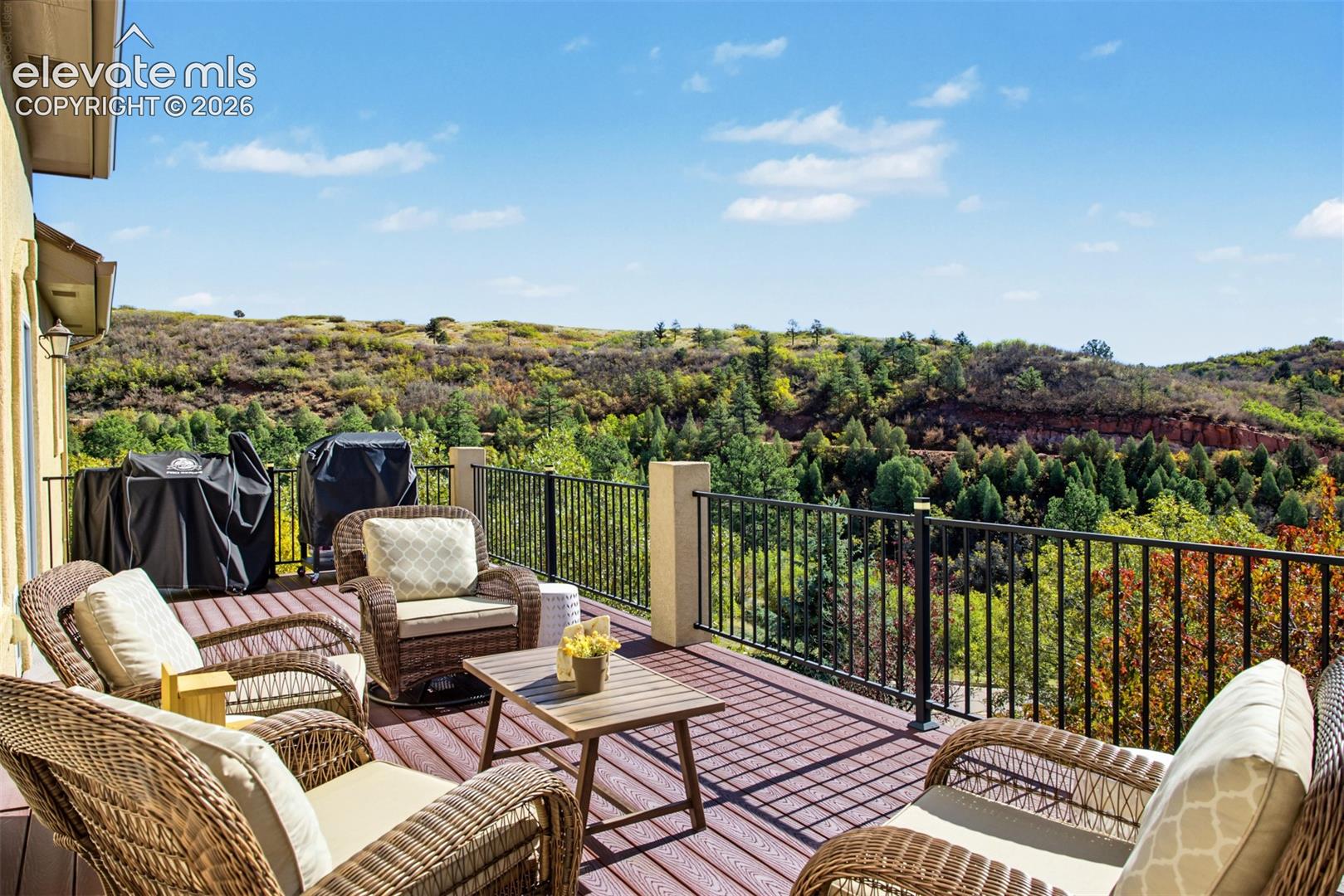 Spacious back balcony offering an inviting outdoor living and entertaining space with sweeping views of the surrounding foothills, mature trees, and natural landscape. This angle faces directly toward Garden of the Gods, overlooking the scenic ridgeline with Rampart Range Road in the distance, a historic mountain road winding through Pike National Forest toward Woodland Park.