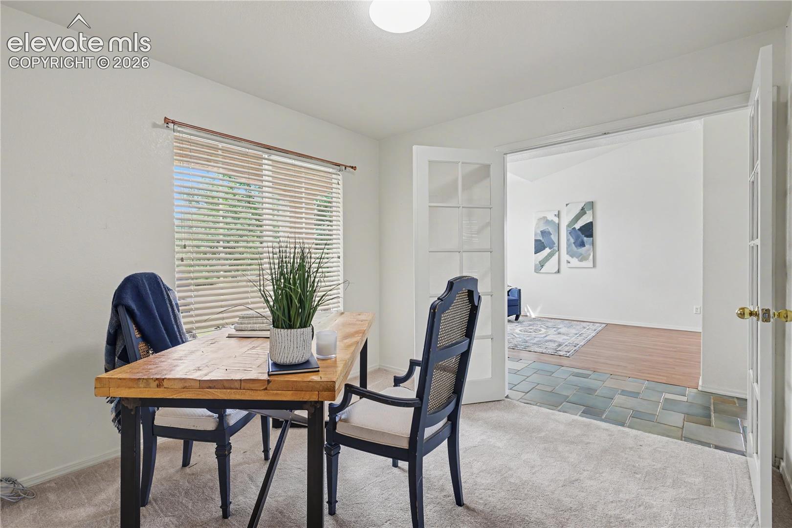 Versatile room featuring light carpeting, a large window with horizontal blinds, and double French doors opening to an additional area with slate tile and wood-finish flooring
