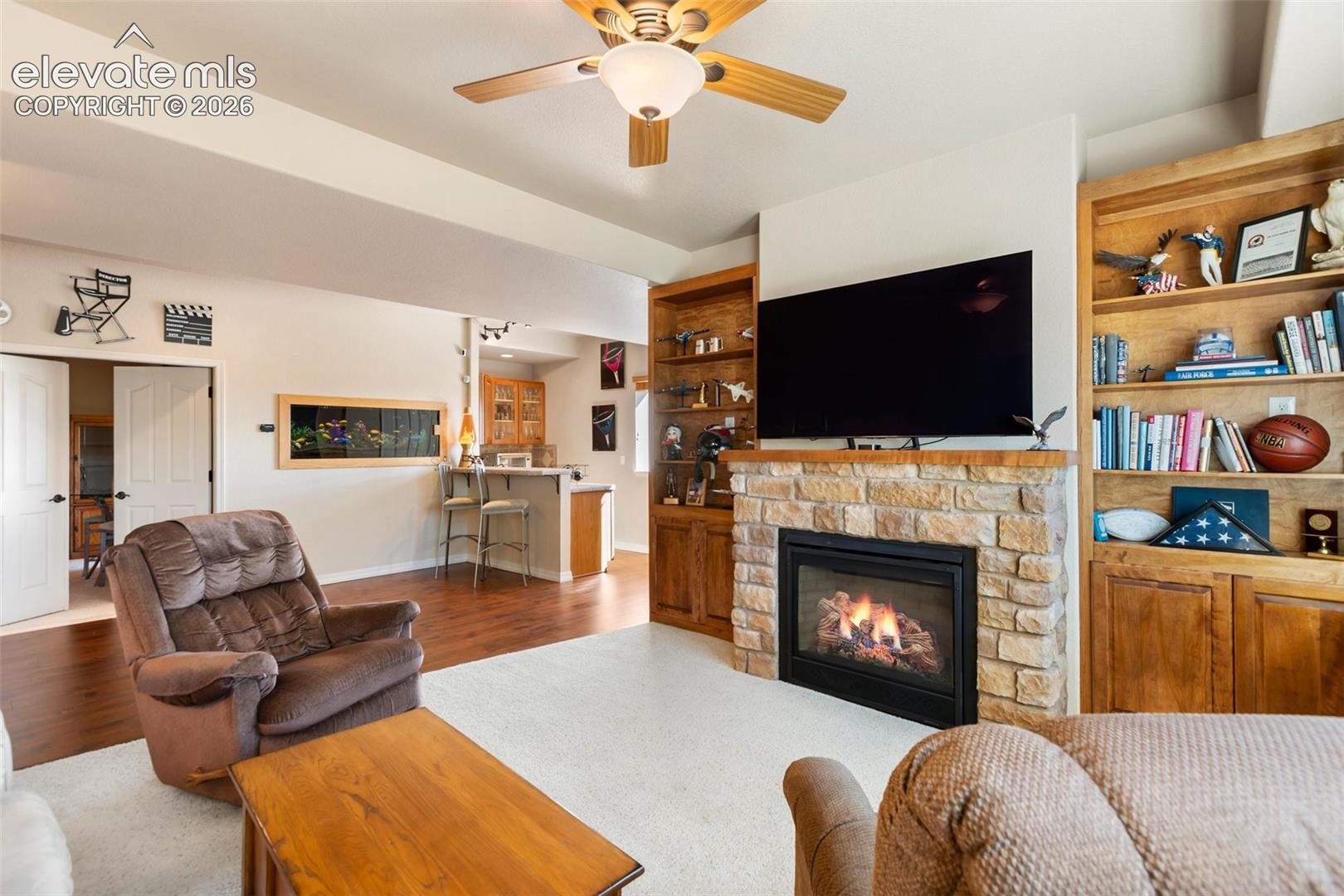 Stone-surround fireplace with a wooden mantel, integrated floor-to-ceiling wooden cabinetry, wood-finish flooring, and a ceiling fan with light fixture