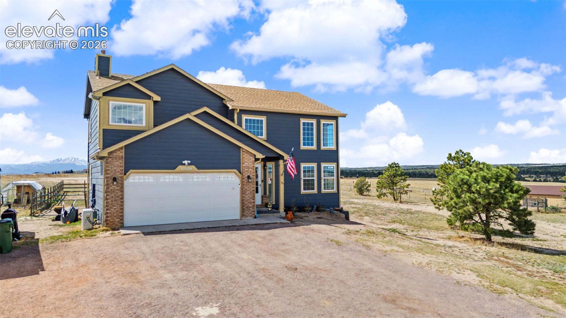 Two-story residence featuring blue horizontal lap siding, brick masonry accents, an attached two-car garage, and a multi-gabled asphalt shingle roof, set against a mountainous horizon