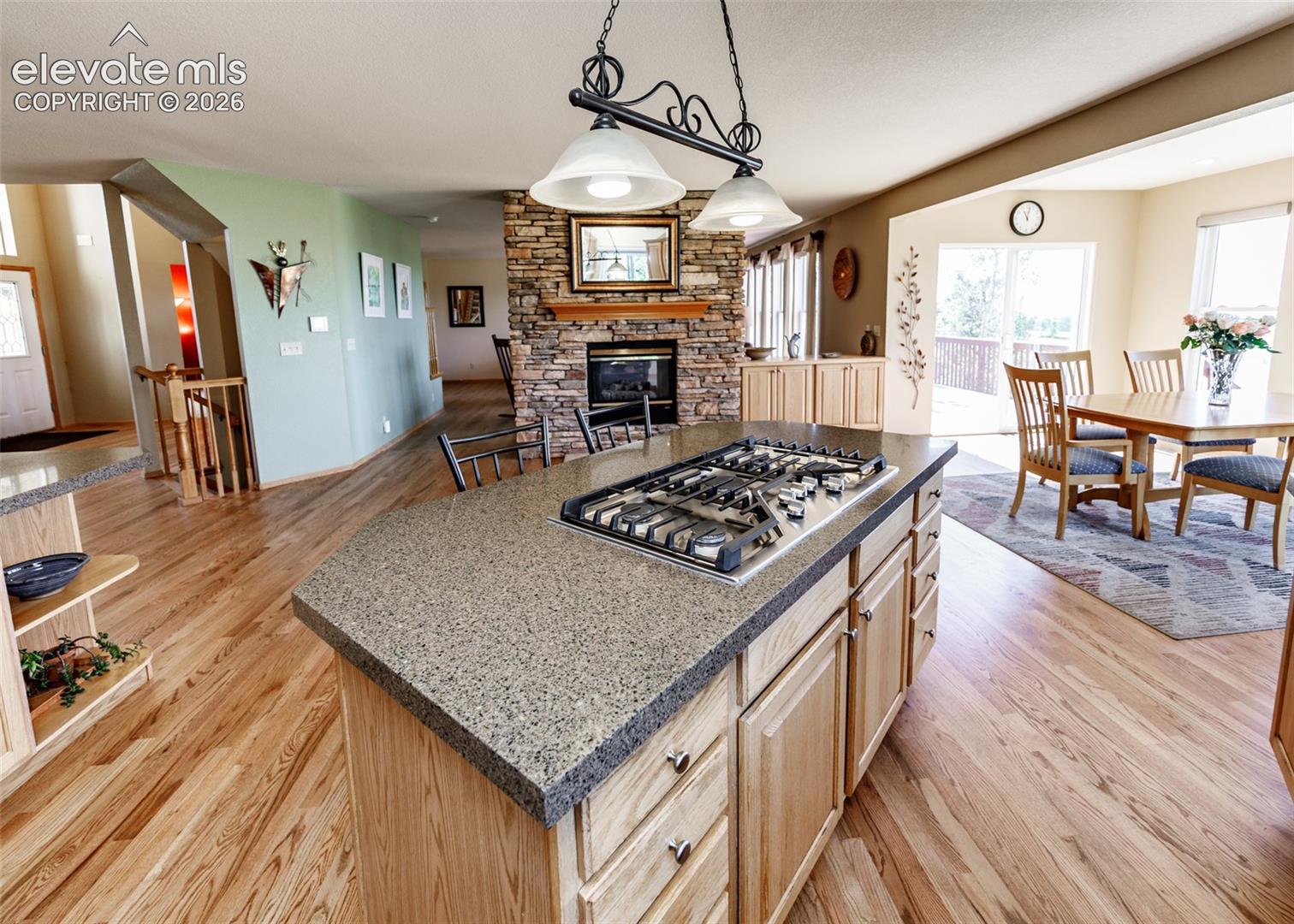 Kitchen island with stone-finish countertop, integrated gas cooktop, wood-finish cabinetry, wood-finish flooring, and floor-to-ceiling stone-veneer fireplace