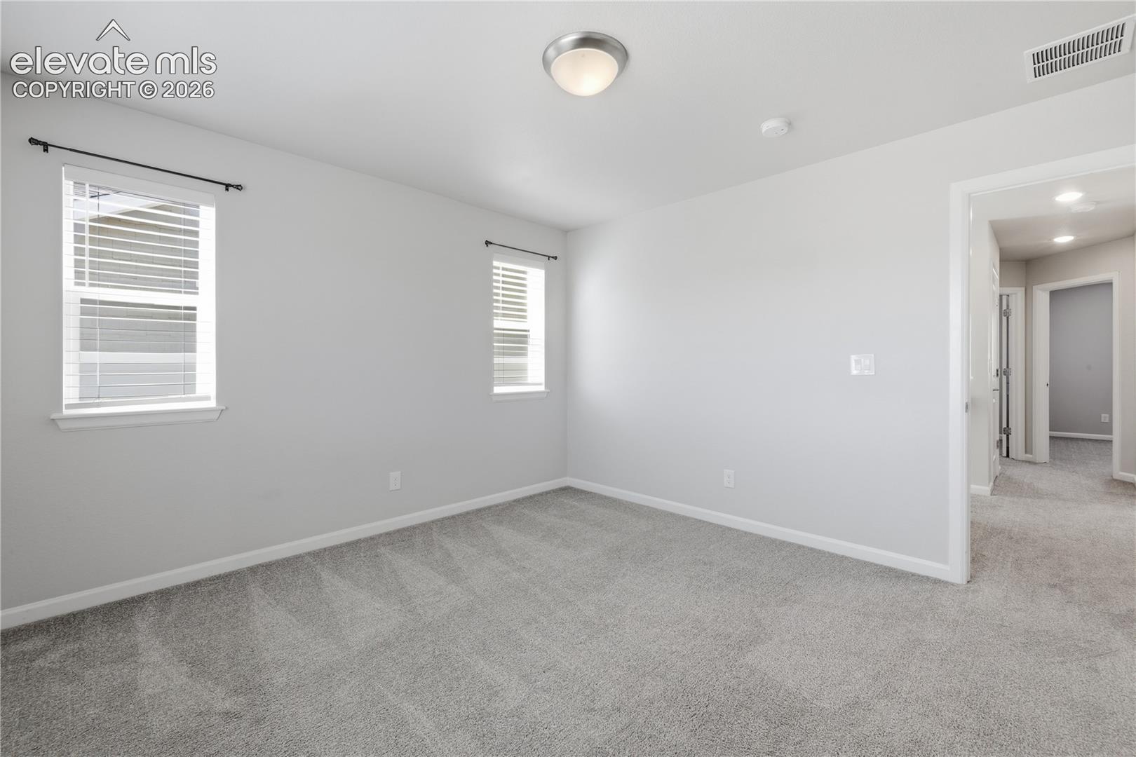 Spacious interior room featuring plush gray carpeting, neutral wall finishes, two windows with blinds, a ceiling-mounted light fixture, and white baseboards
