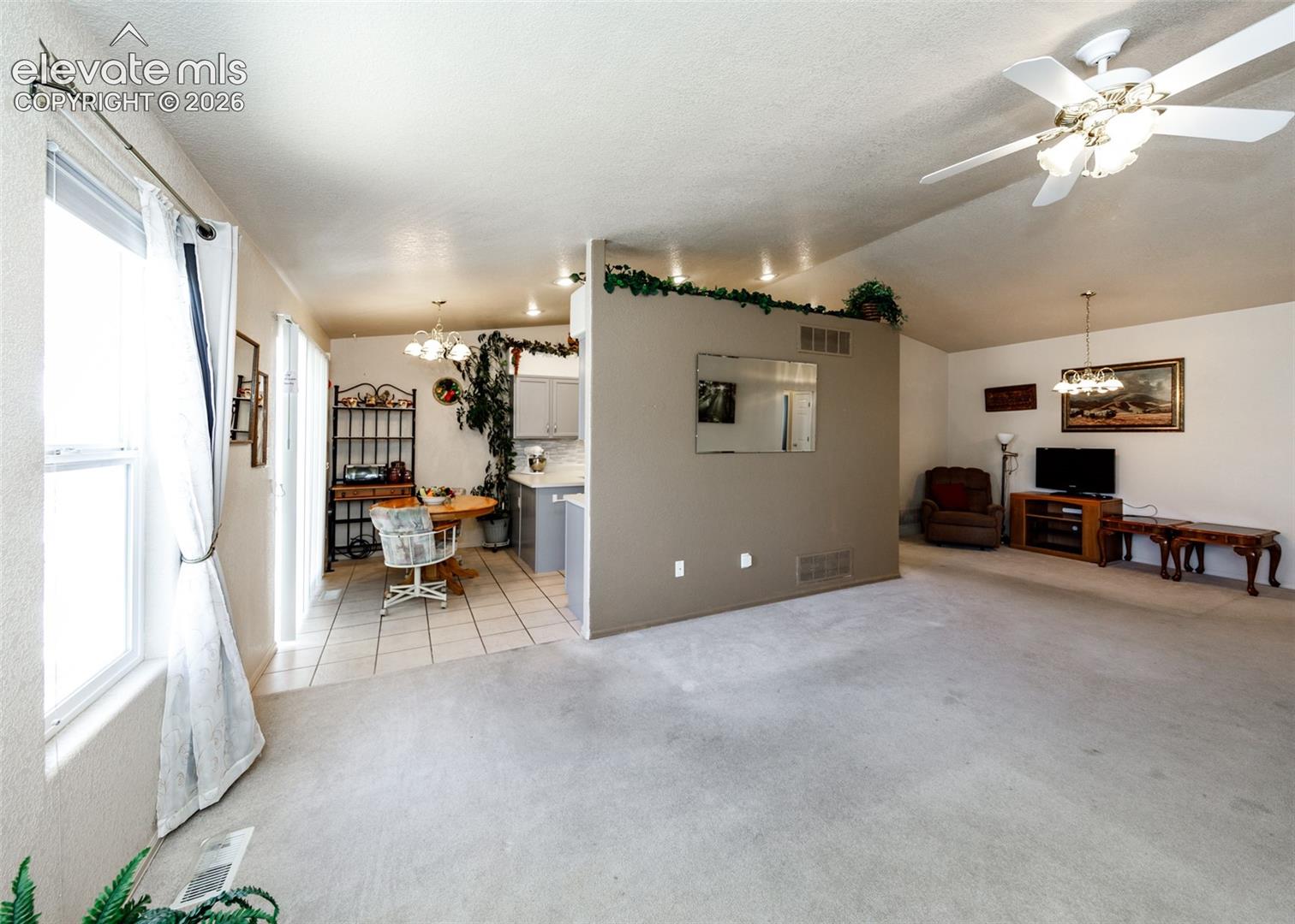 Living area featuring vaulted ceilings, central ceiling fan, neutral wall-to-wall carpeting, tile flooring in the kitchen area, and a centrally positioned partition wall