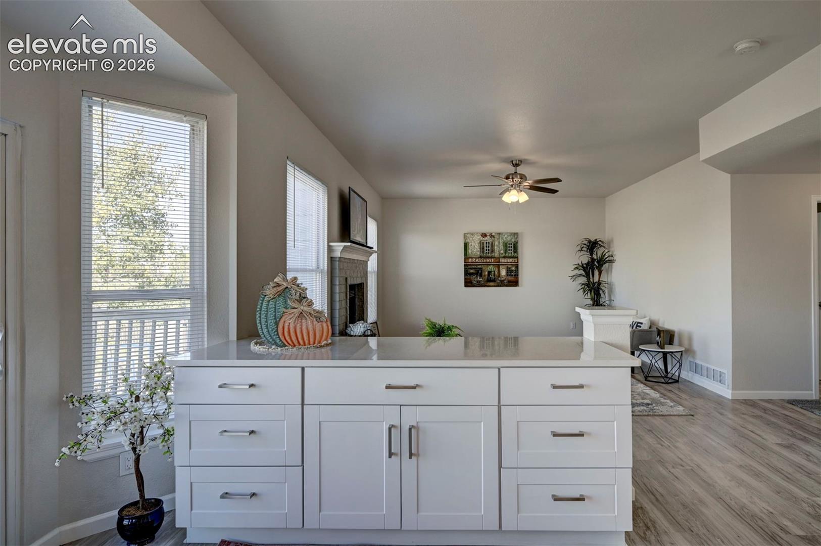 Kitchen island featuring white cabinetry, metallic hardware, and a solid-surface countertop, complemented by wood-finish flooring, multiple windows with blinds, and a ceiling-mounted light fixture