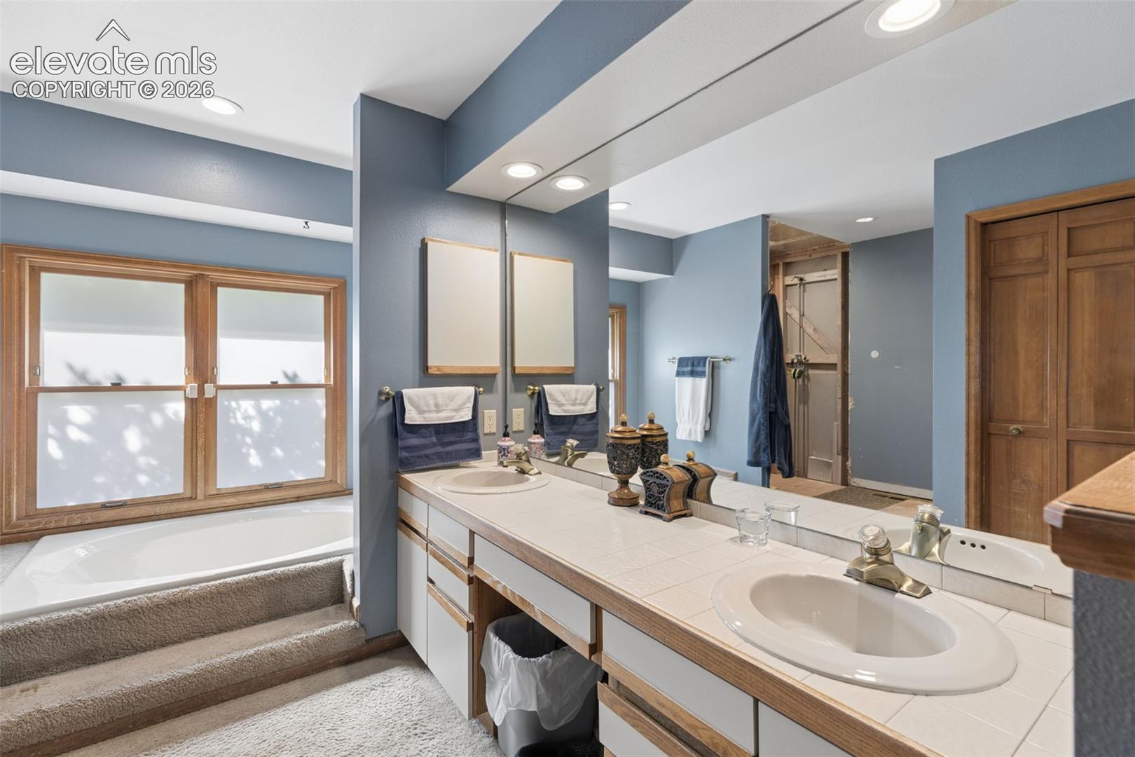 Dual-sink vanity with tile countertop, integrated soaking tub, wood-trimmed window, and full-width mirror