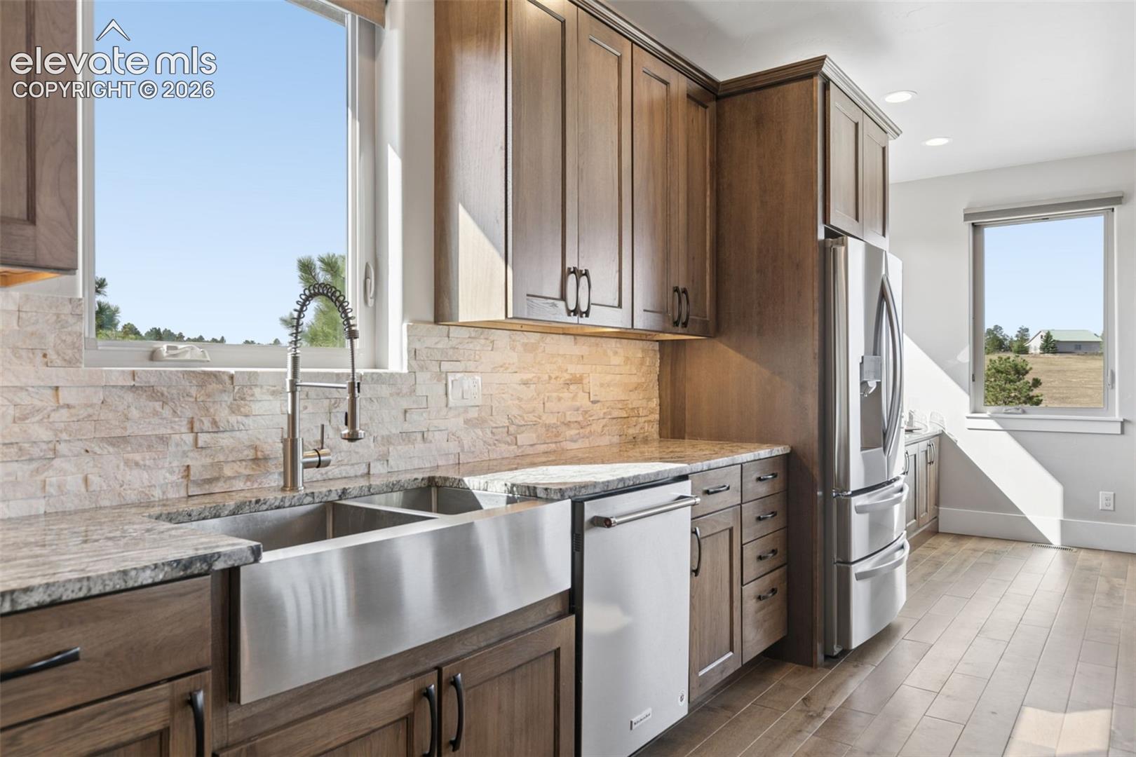 Wood-finish cabinetry, stainless steel farmhouse sink, stacked stone backsplash, light-toned countertops, and wood-finish flooring