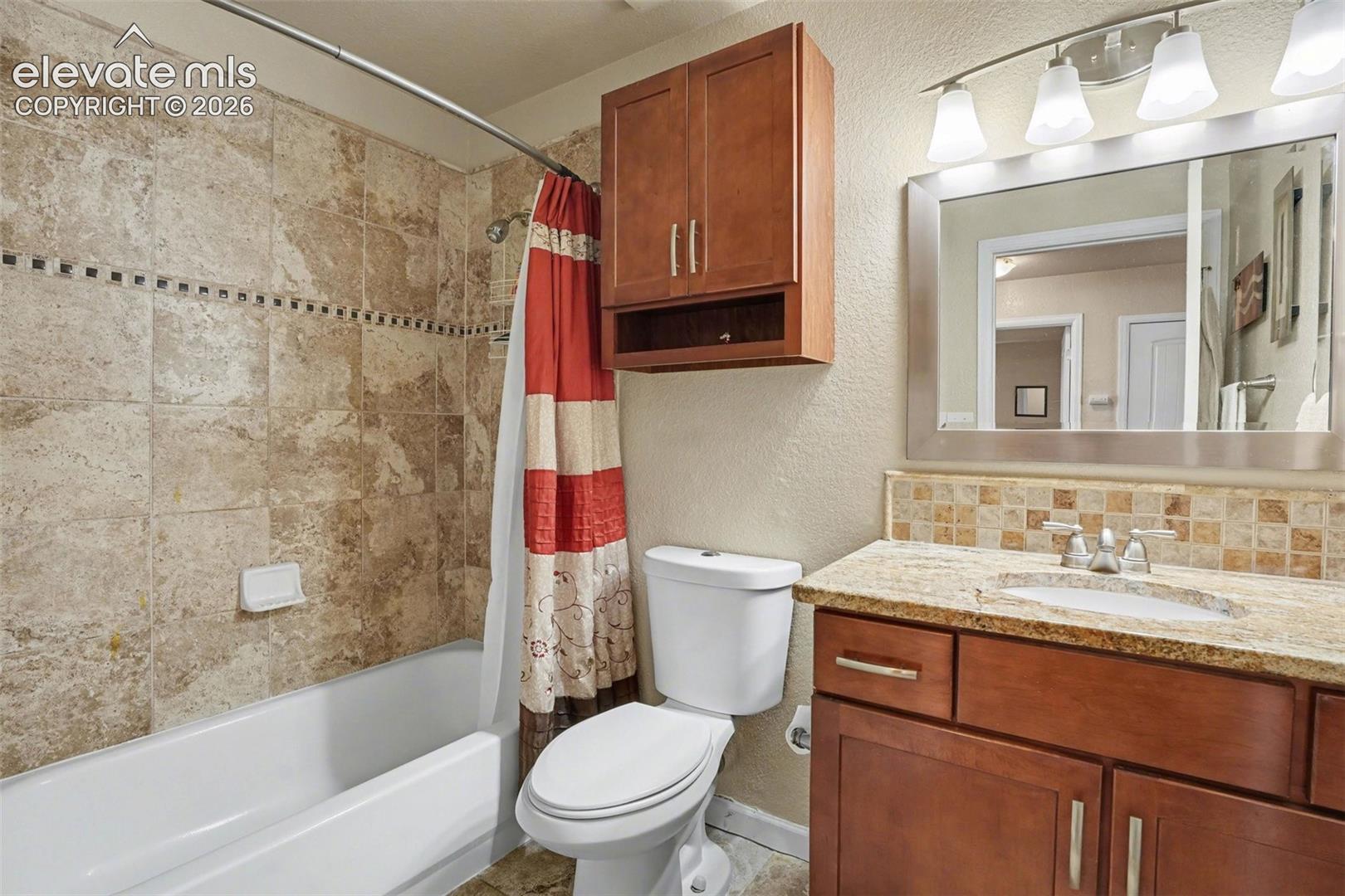 Bathroom featuring a stone-tile shower surround with decorative inlay, wood-finish cabinetry, a stone-surfaced vanity with an integrated sink, and an expansive wall-mounted mirror