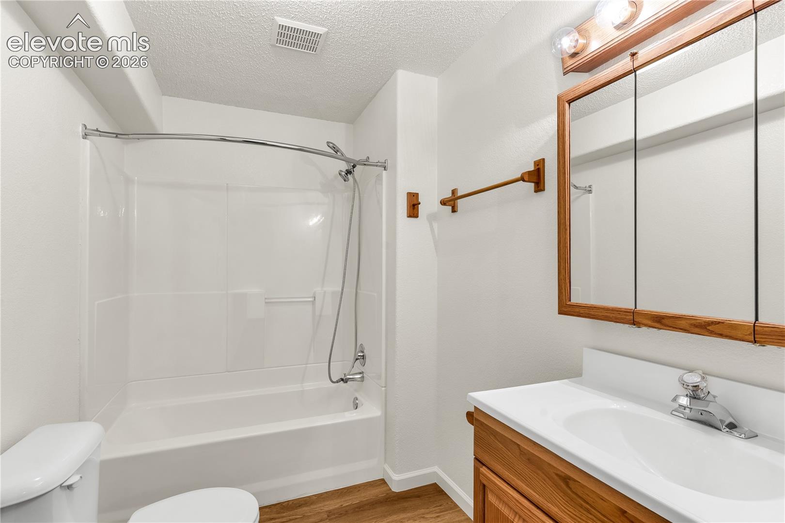 Bathroom featuring wood-finish flooring, a white integrated sink with laminate countertop, a wood-framed medicine cabinet, a full-size bathtub with white surround, and a curved shower rod