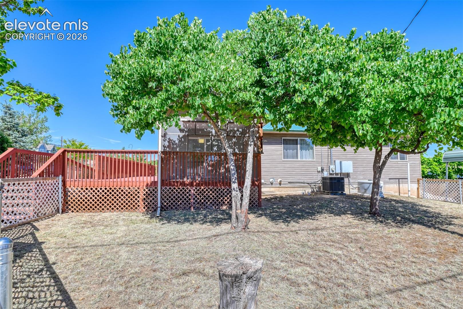 Backyard featuring a raised wooden deck with lattice skirting, metal fencing, and mature trees