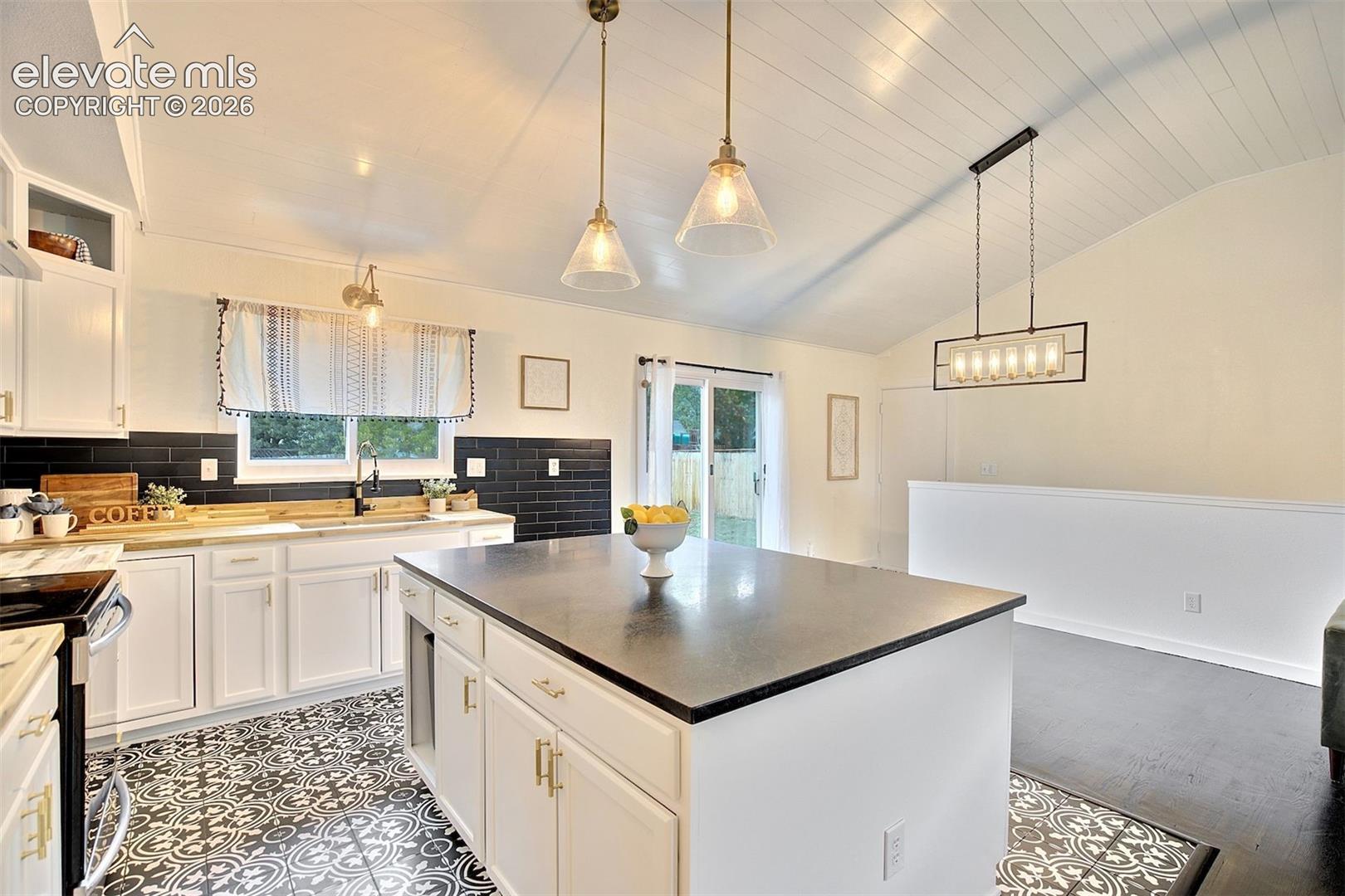 Kitchen featuring a central island with a dark countertop, white cabinetry with gold-tone hardware, patterned tile flooring, black subway tile backsplash, and vaulted ceilings with pendant lighting