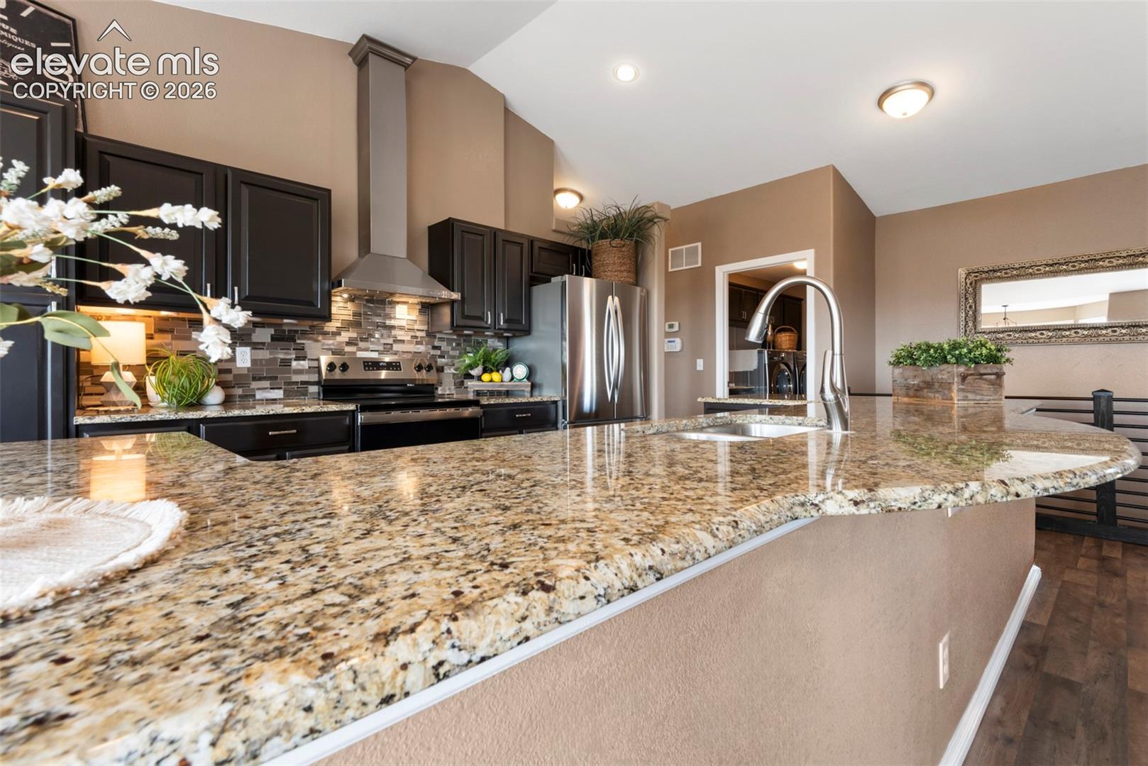 Kitchen featuring dark cabinetry, stone countertops, stainless steel chimney-style range hood, multi-tone tile backsplash, and wood-finish flooring