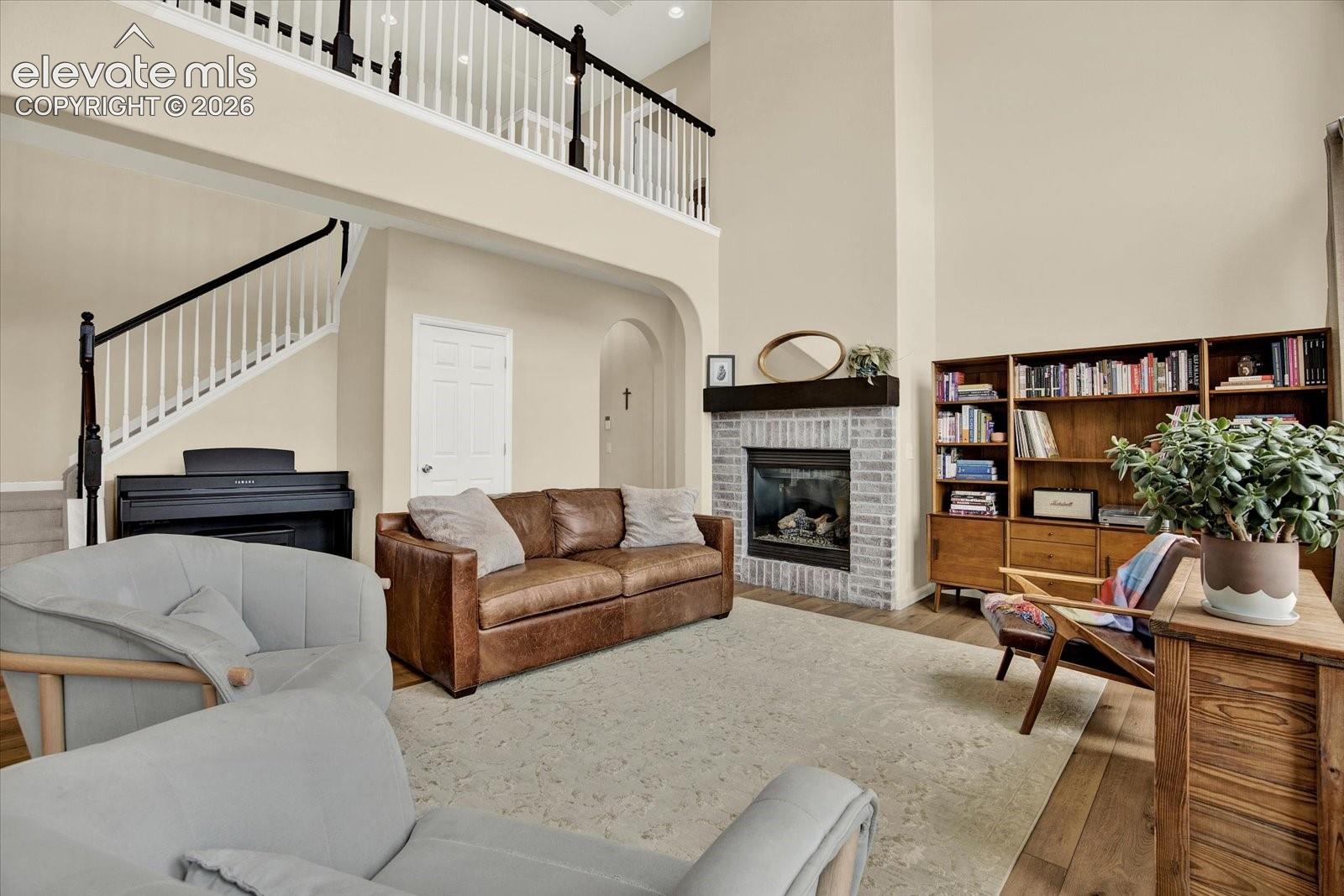 Double-height ceiling, brick-surround fireplace with a wooden mantel, built-in wooden shelving, staircase with a custom spindle railing, and wood-finish flooring