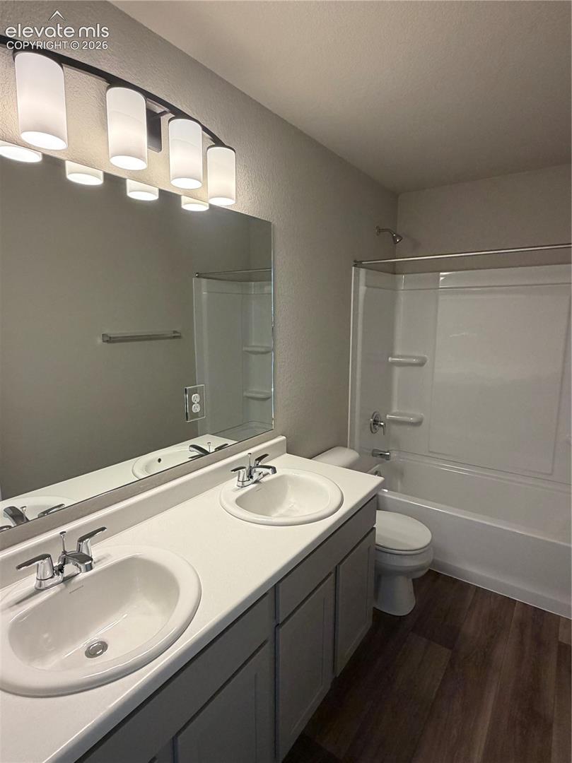 Dual vanity with white solid-surface countertop, drop-in sinks, chrome fixtures, full-width mirror with multi-light fixture, and wood-finish flooring