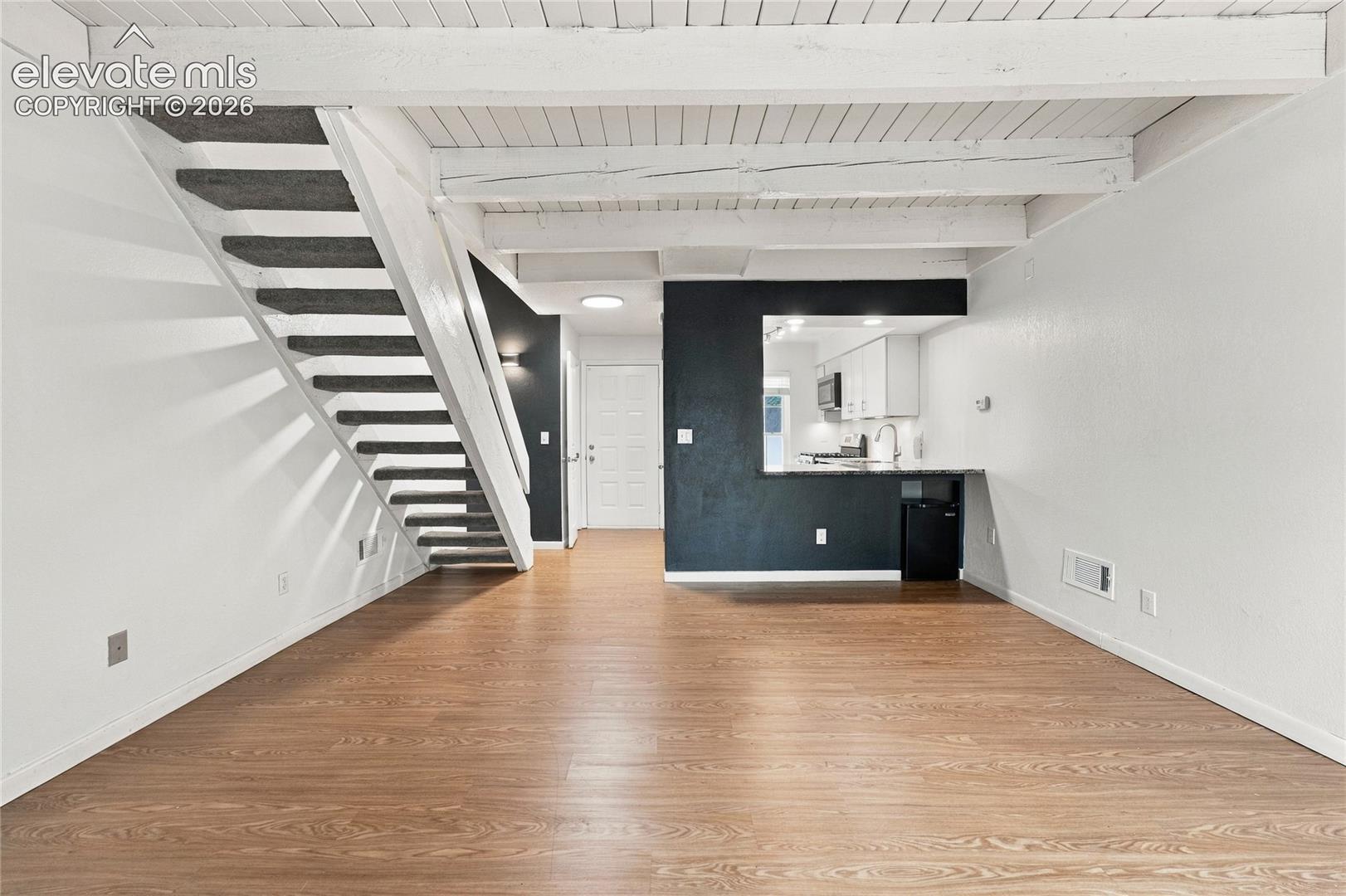 Open-concept interior featuring exposed white-painted ceiling beams, wood-finish flooring, a staircase with carpeted treads, and a kitchen pass-through window with a stone-surface breakfast bar