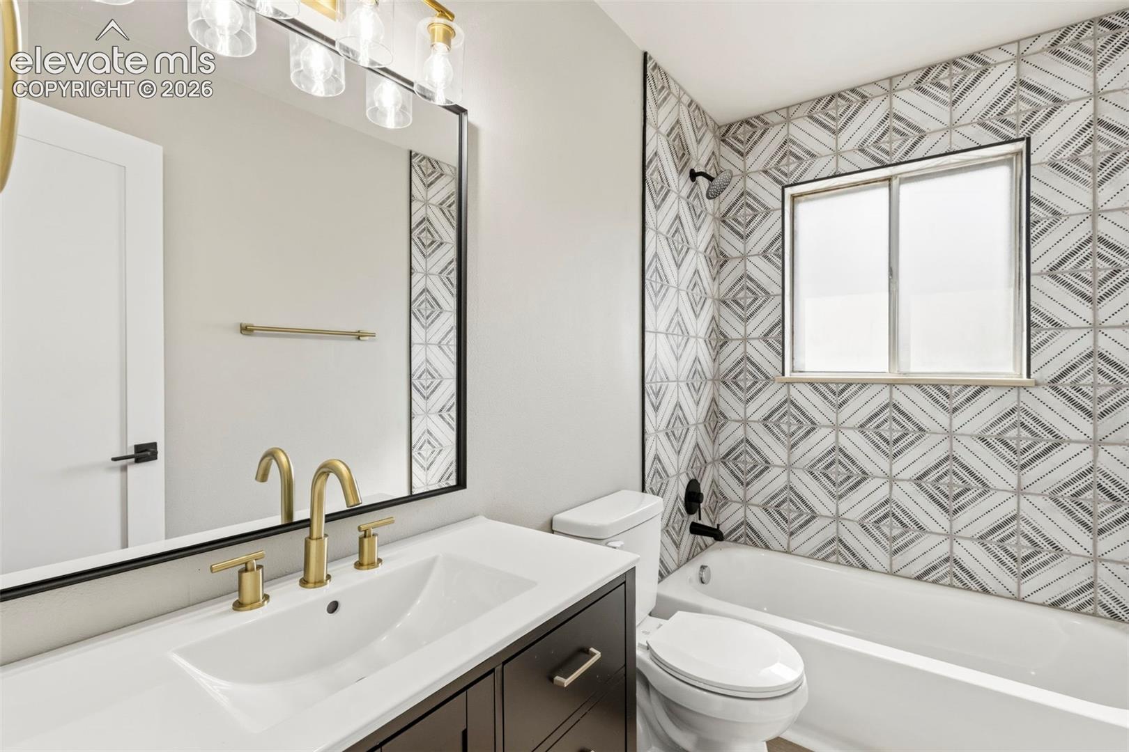 Bathroom featuring a dark wood-finish vanity with a white countertop and undermount sink, gold fixtures, a rectangular framed mirror, and a wall-mounted light fixture with clear glass shades