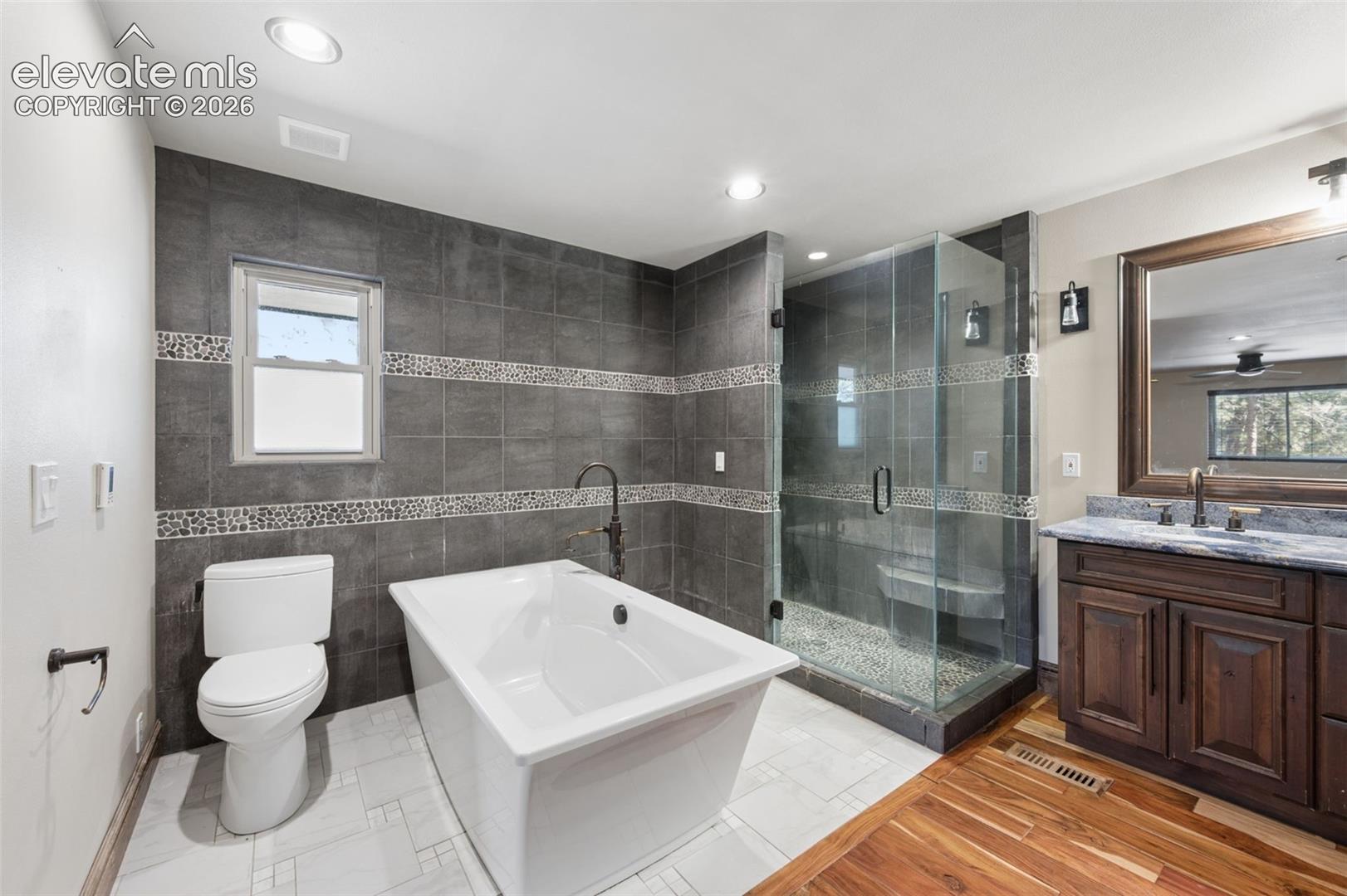 Freestanding soaking tub with a wall-mounted faucet, frameless glass-enclosed shower with tiled seating, dark wood-finish cabinetry, stone countertop with an integrated sink, and wood-finish flooring