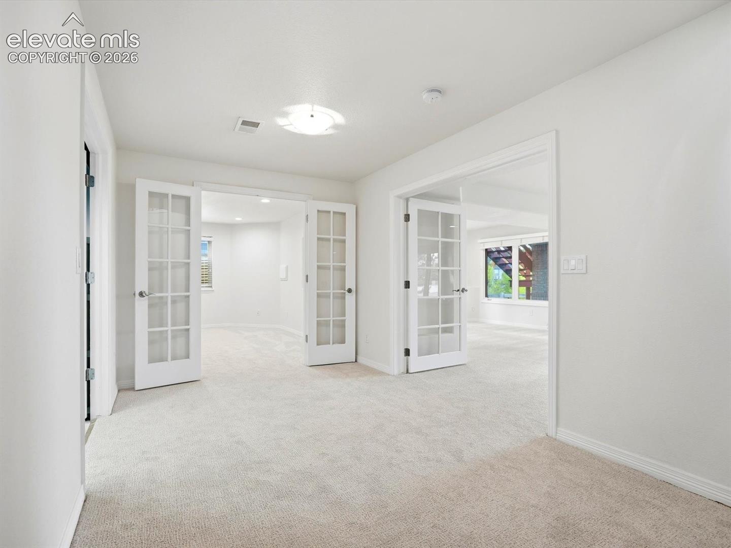 Neutral-toned wall-to-wall carpeting and crisp white interior paint, highlighted by two sets of double French doors with glass panes and flush-mount ceiling lighting