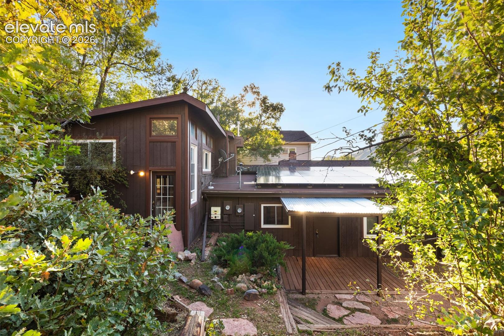 Exterior featuring brown wood siding, roof-mounted solar panels, wood-decked patio, and natural stone hardscaping surrounded by mature trees