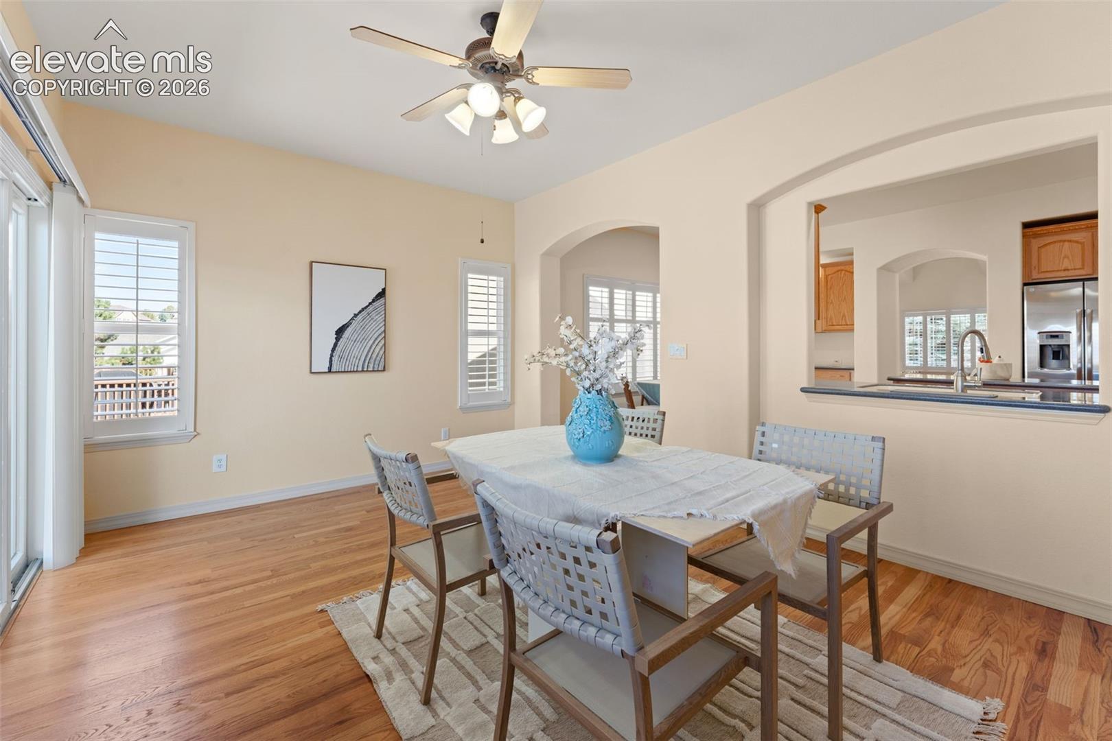 Wood-finish flooring, plantation shutters, arched architectural detailing, ceiling fan with integrated lighting, and pass-through counter