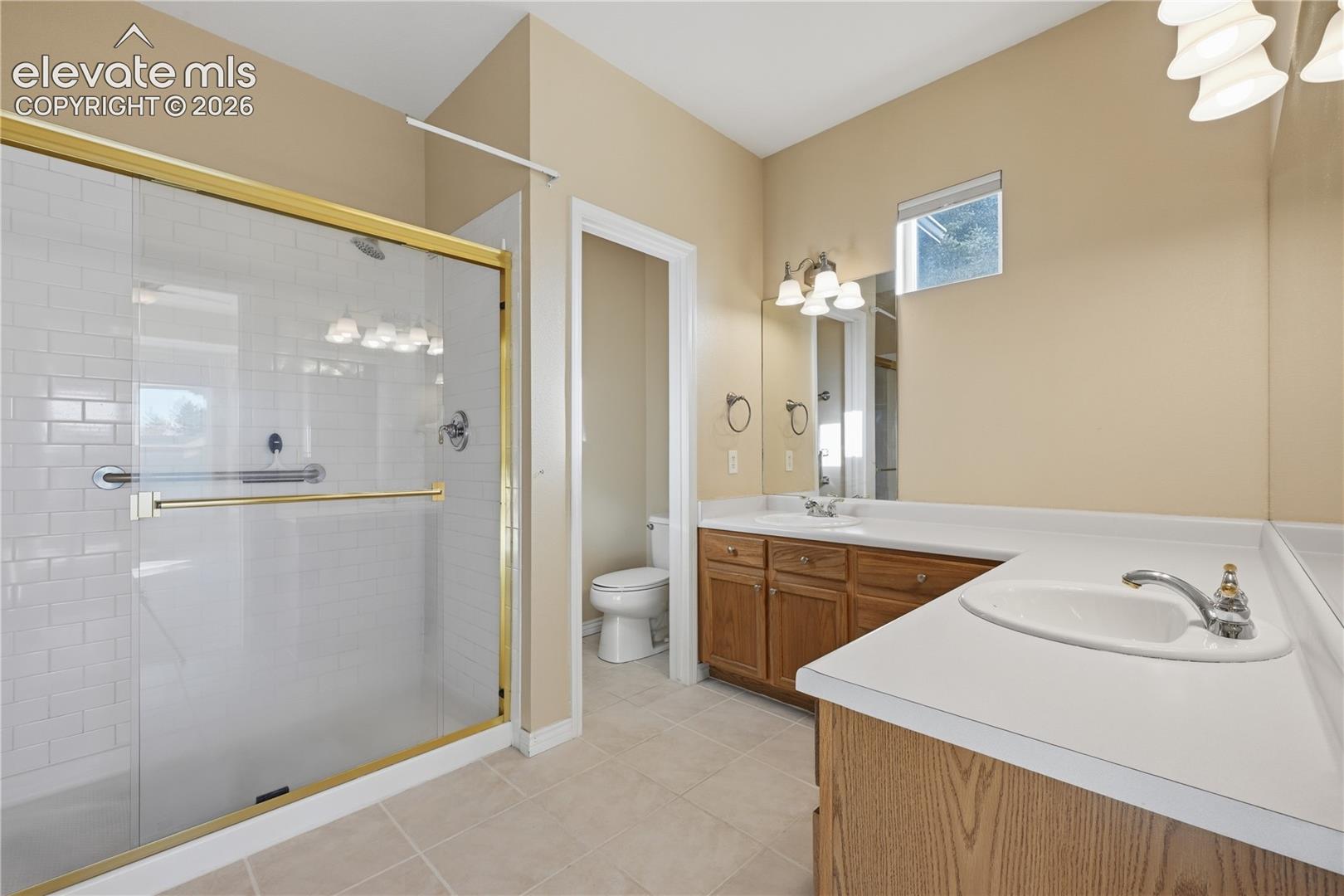 Bathroom featuring a dual-sink vanity with wood cabinetry and solid-surface countertops, a walk-in shower with white subway-style tile, a brass-finish sliding glass enclosure, and neutral tile flooring
