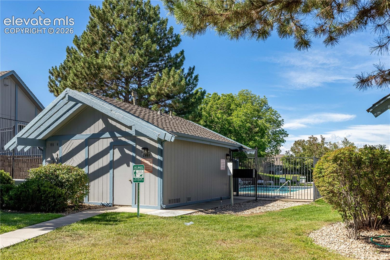 Community building with wood-paneled siding, asphalt shingle roof, attached wall-mounted lighting, and nearby swimming pool enclosed by metal fencing