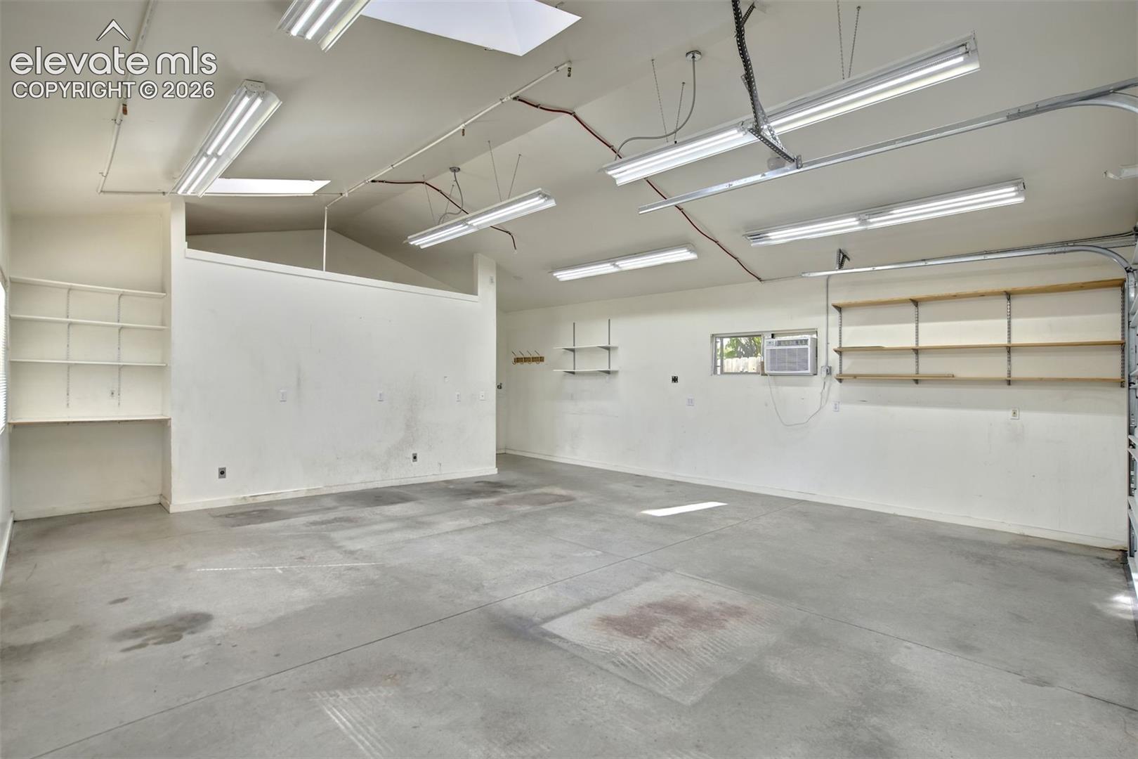 Spacious garage featuring vaulted ceilings, linear fluorescent lighting, built-in wall shelving, and a wall-mounted climate control unit