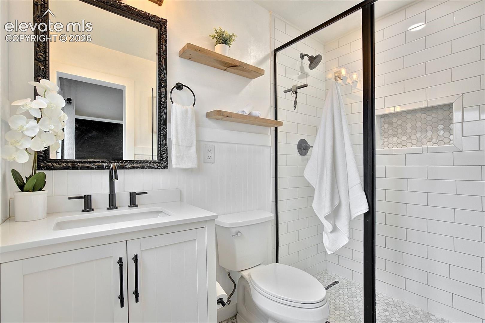 Bathroom featuring a white vanity with a rectangular sink, matte black fixtures, subway-tiled walk-in shower with a glass enclosure, built-in wall niche, and natural-finish floating wood shelving
