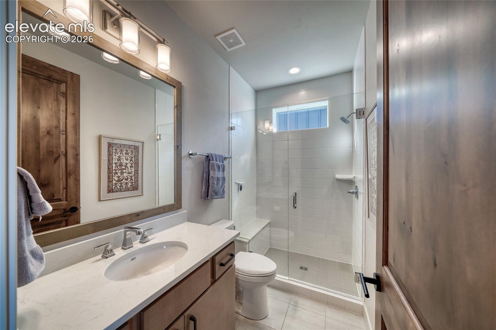 Bathroom featuring a vanity with a light-toned countertop, a wood-framed mirror, a glass-enclosed shower with tiled walls and a built-in bench, and wood-finish doors