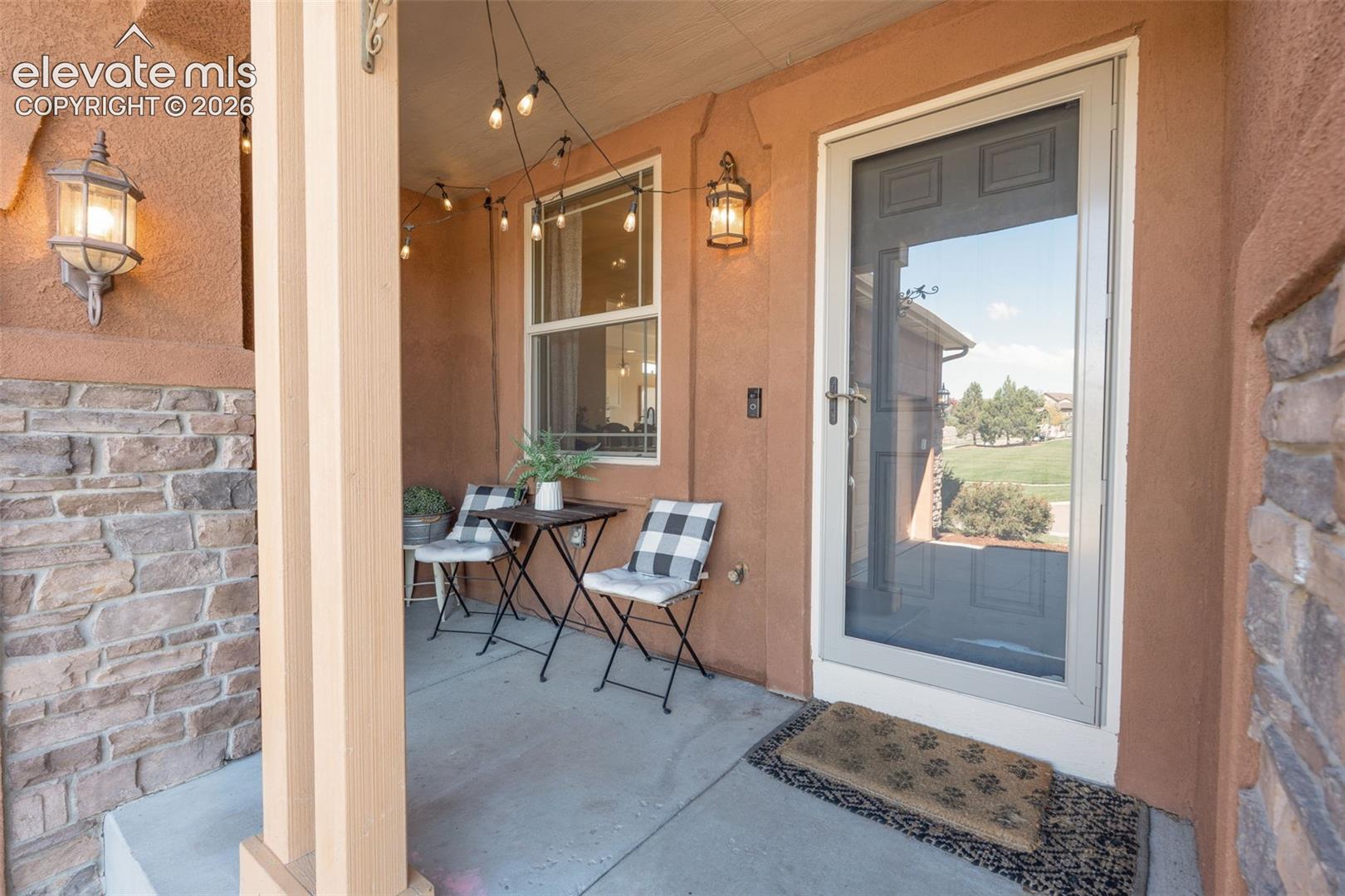 Covered front porch featuring stucco walls, stone veneer accents, concrete flooring, and two wall-mounted lanterns