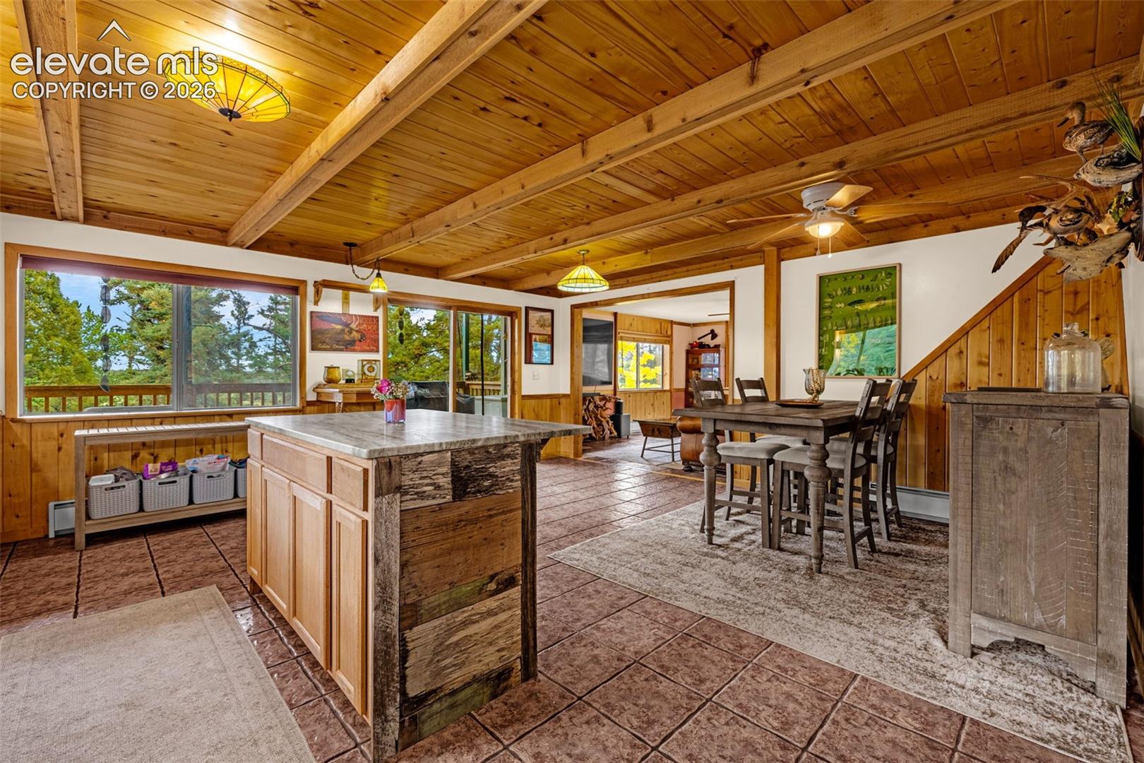 Exposed beam ceiling with wood paneling, central kitchen island with stone countertop and wood-paneled exterior, tile flooring, and sliding glass doors