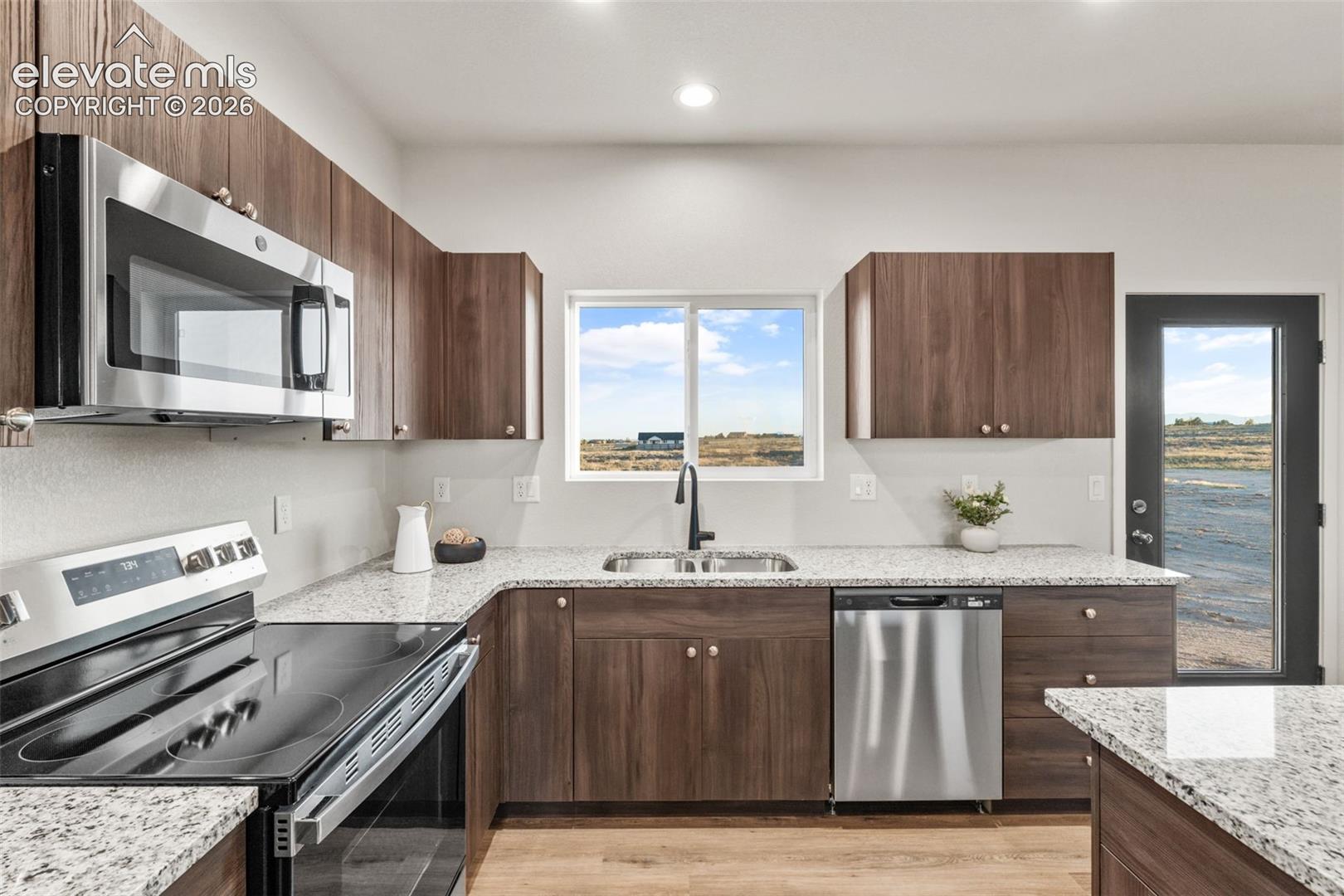 Kitchen featuring dark-toned flat-panel cabinetry, light-colored speckled countertops, stainless steel appliances, wood-finish flooring, and a single-handle matte black faucet