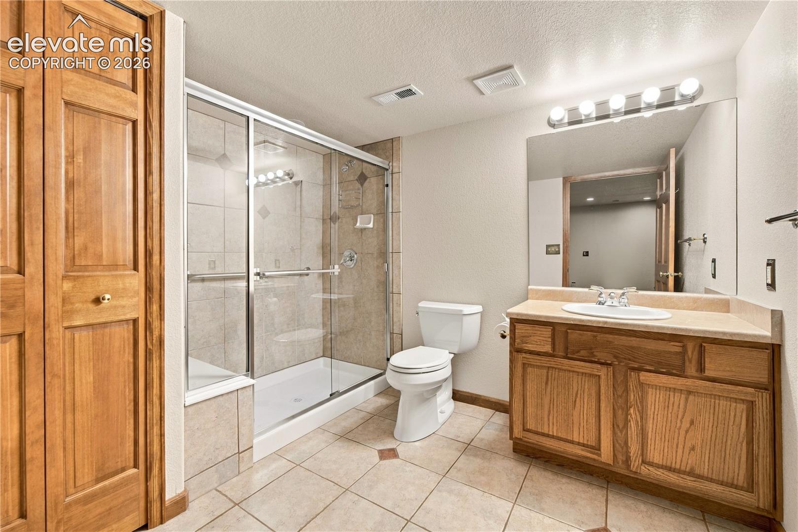 Bathroom featuring wood-finish cabinetry, a glass-enclosed shower with tile surround, neutral tile flooring, and a full-width vanity mirror with integrated lighting