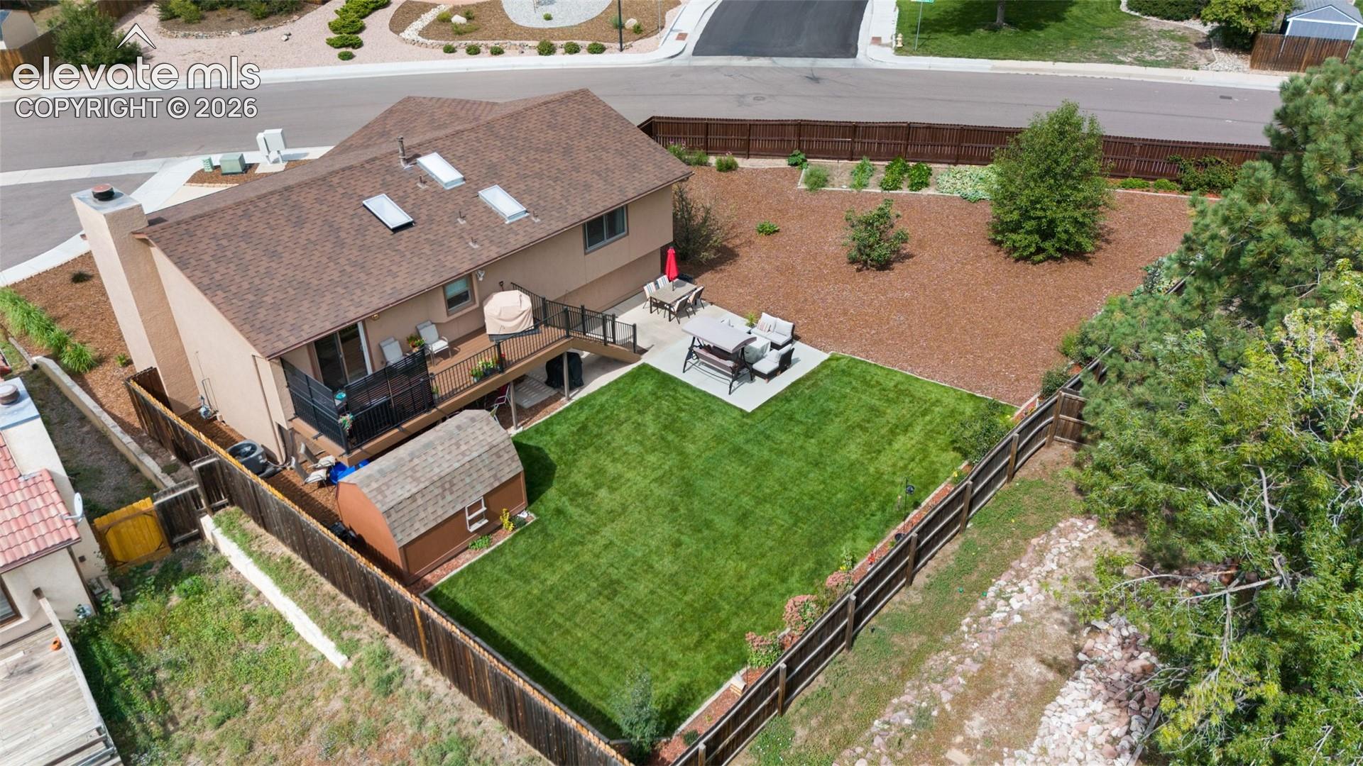 Aerial view of a backyard featuring a raised wooden deck with metal railing, a paved concrete patio, a manicured lawn, and a fenced perimeter