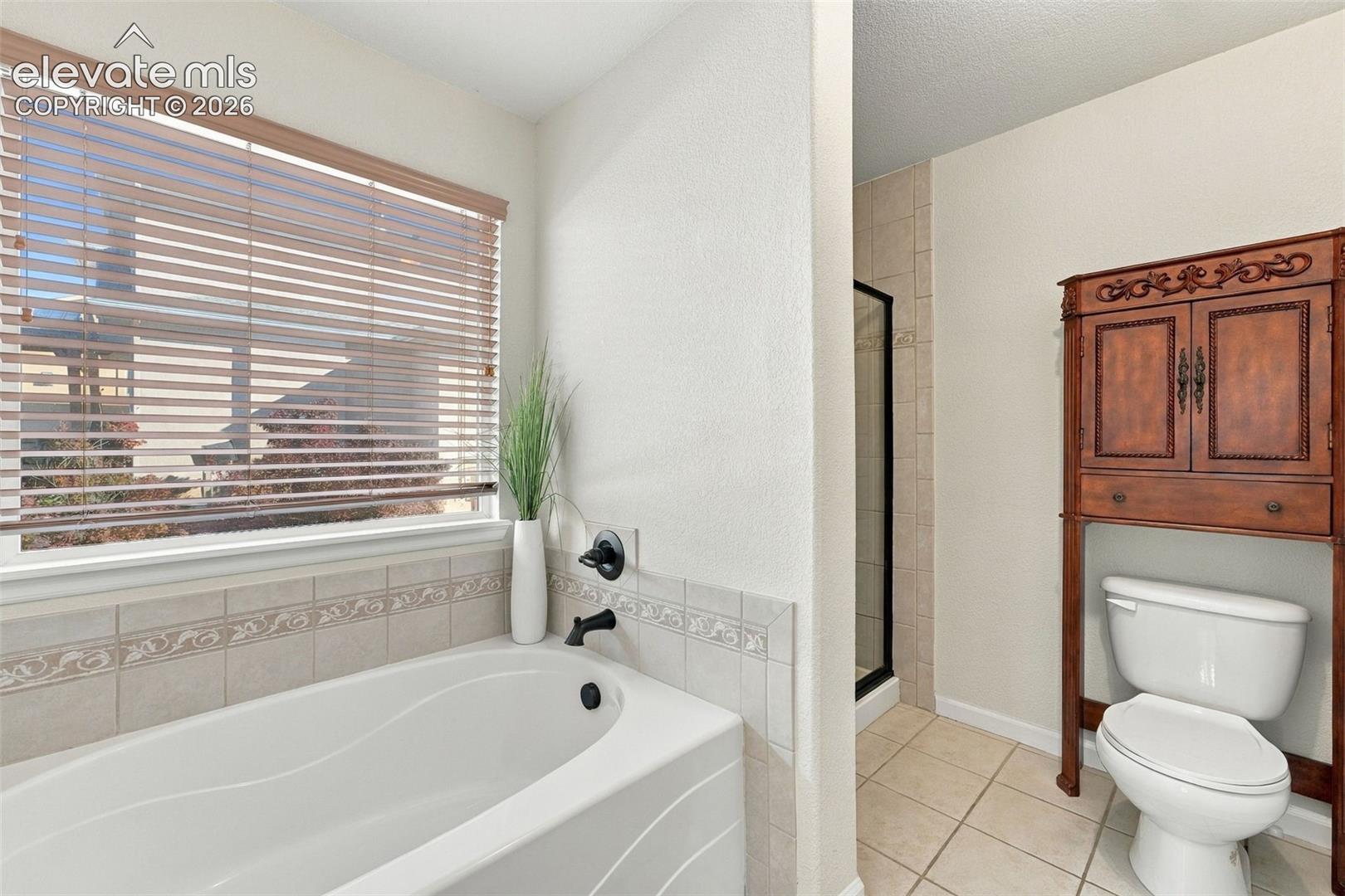 Deep soaking tub with matte black fixtures, ceramic tile surround with decorative inlay, horizontal blind window treatment, and separate enclosed shower stall with tiled interior
