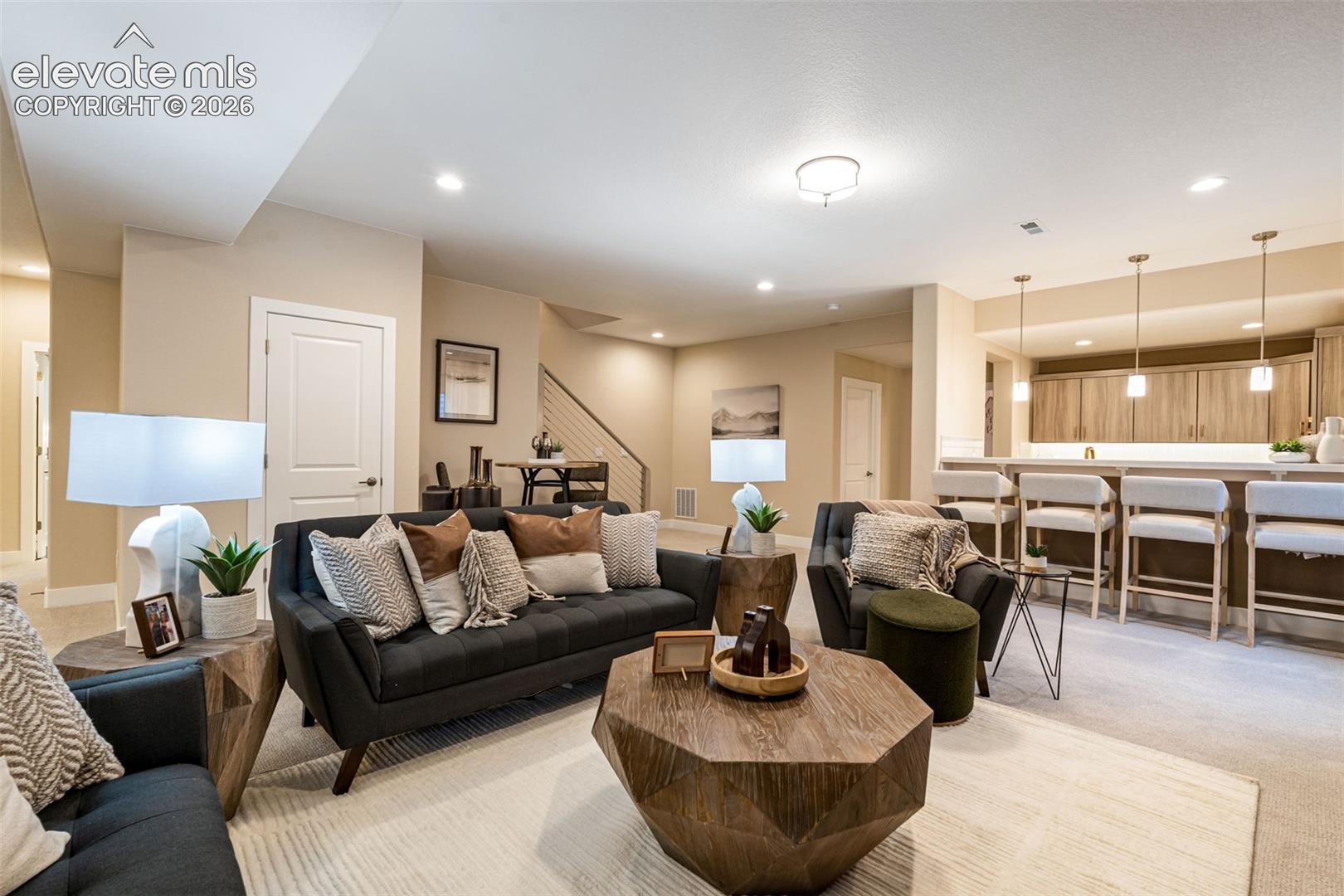 Open-concept recreation area featuring a wet bar with pendant lighting, neutral wall finishes, recessed lighting, and a metal-railing staircase