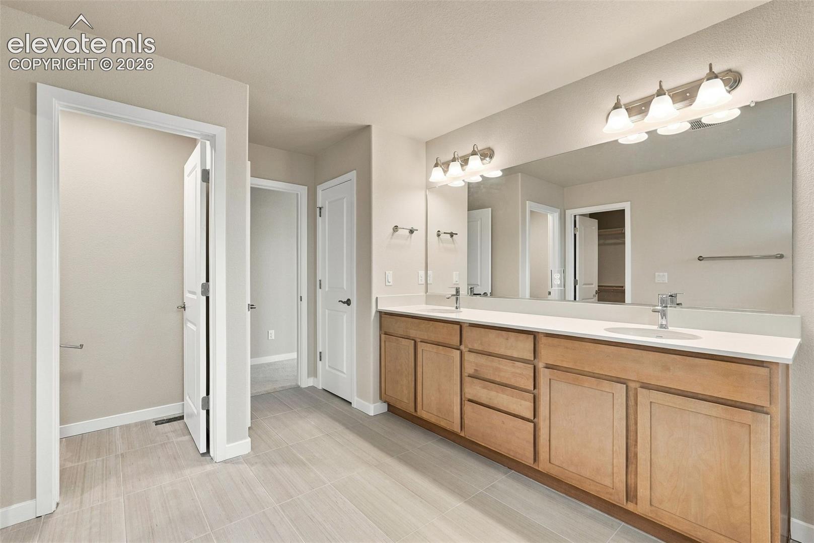 Dual-sink vanity with light wood cabinetry, white countertops, two wall-mounted light fixtures, and oversized frameless mirror