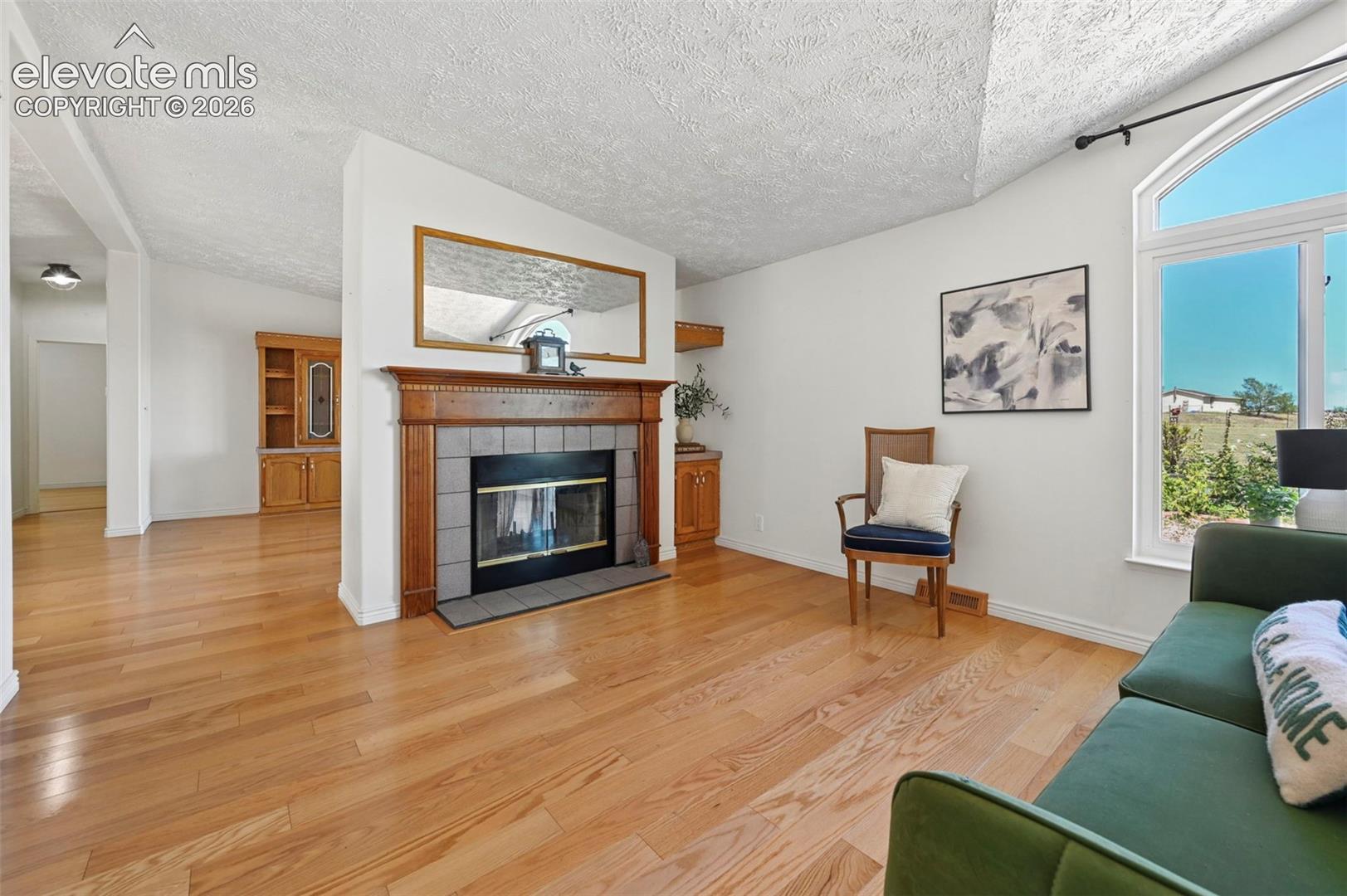Hardwood flooring throughout, a fireplace with a wood mantel and tile surround, built-in shelving, and an arched window