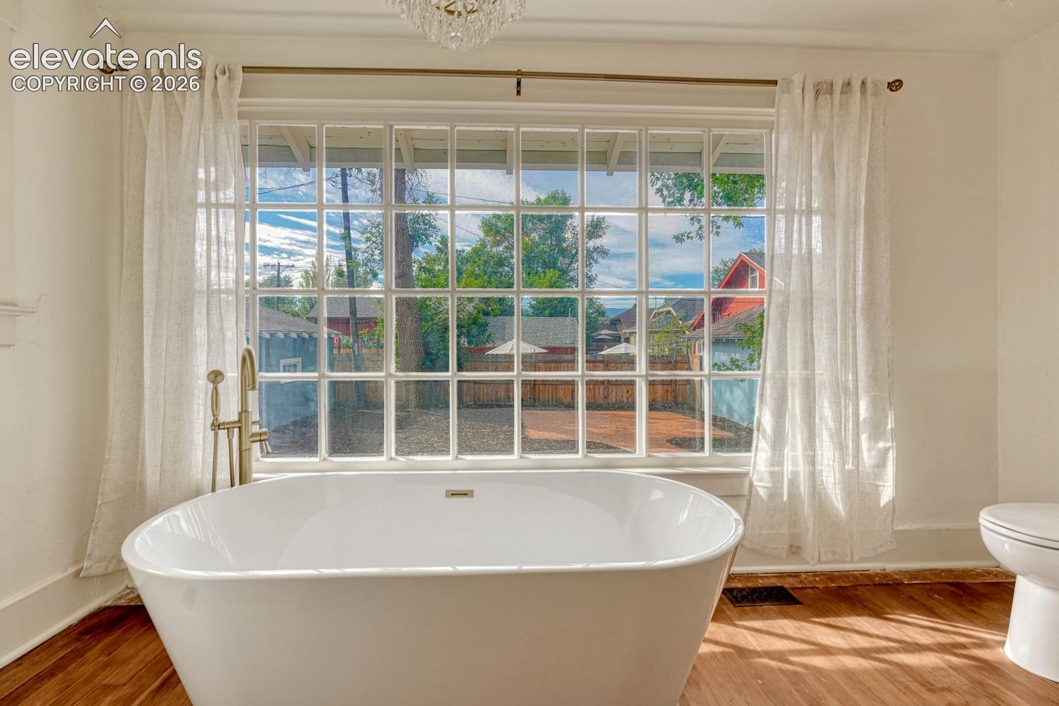 Freestanding soaking tub with a floor-mounted brass fixture, large grid-pattern window, and wood-finish flooring