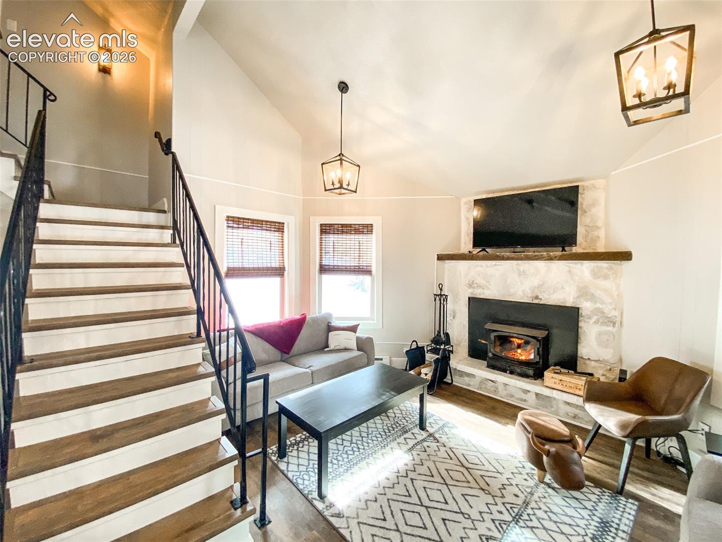 Living room featuring a stone fireplace with a wood-burning stove insert, wood-finish flooring, and a vaulted ceiling with a pendant light fixture