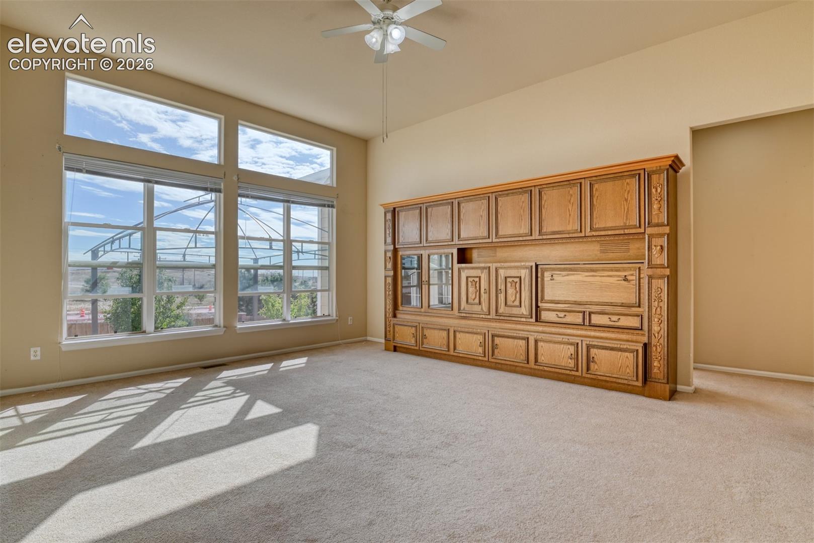 Large windows with horizontal blinds, built-in wooden cabinetry with glass-front shelving, high ceilings, and a ceiling fan