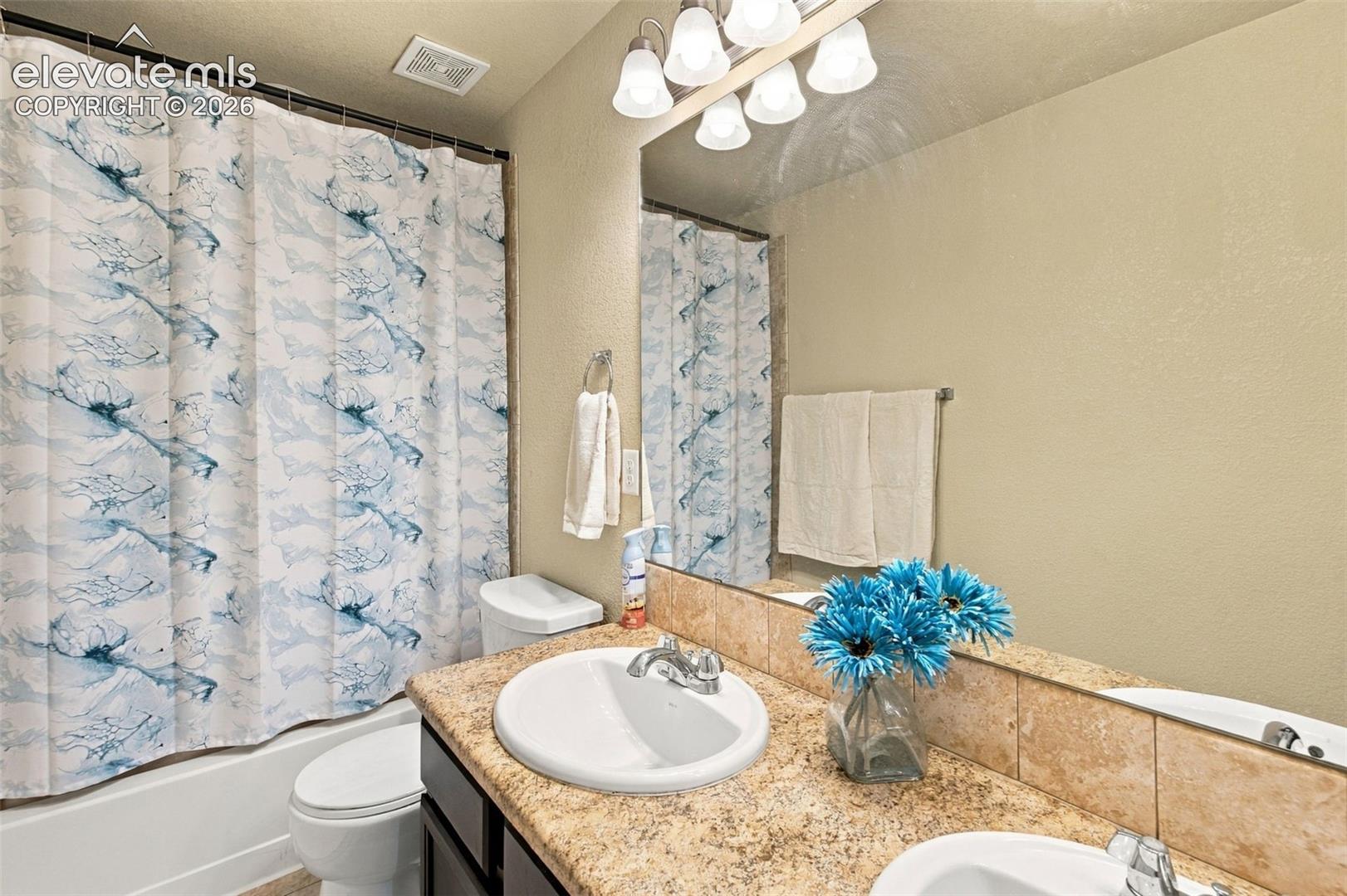 Bathroom featuring a dual-sink vanity with stone-surfaced countertop, full-width wall mirror, five-light overhead fixture, and a tub-shower combination