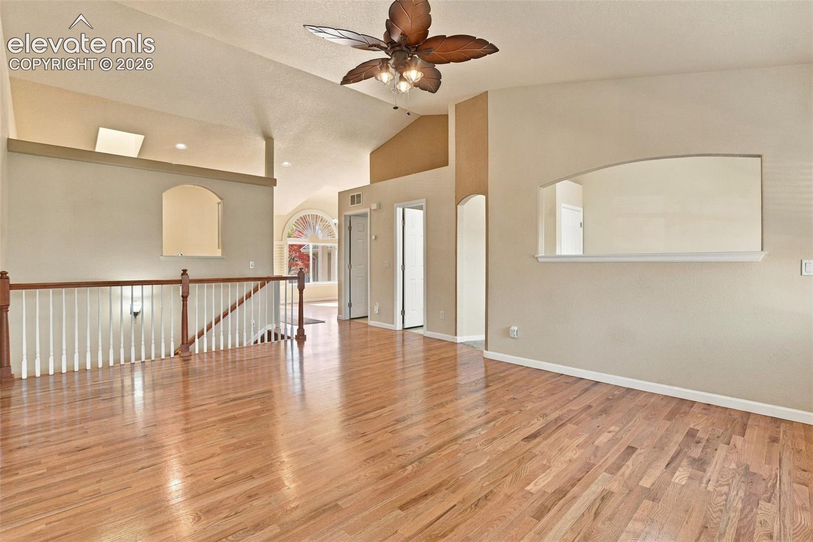 Vaulted ceilings, wood-finish flooring, architectural arched wall niches, decorative ceiling fan with integrated lighting, and white baluster railing