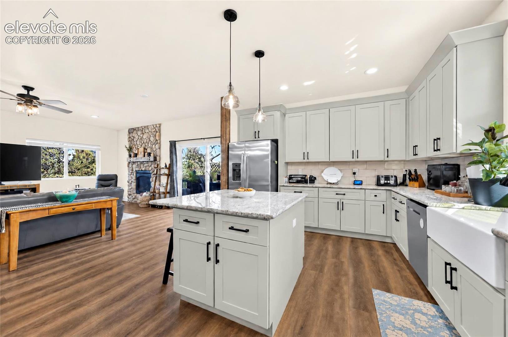 Kitchen featuring light-toned cabinetry, stone-finish countertops, stainless steel appliances, a white farmhouse sink, and a stone-clad fireplace