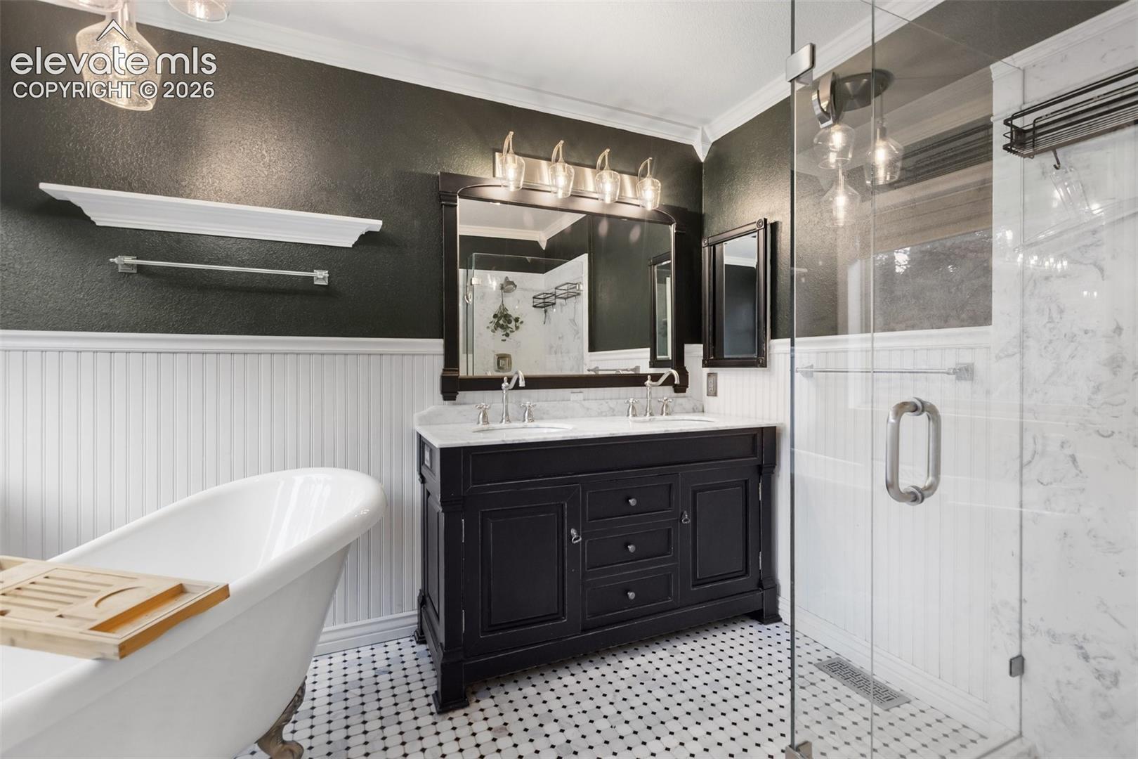 Double vanity with a stone countertop, freestanding soaking tub, walk-in glass-enclosed shower with stone-tile surround, beadboard wainscoting, and patterned tile flooring