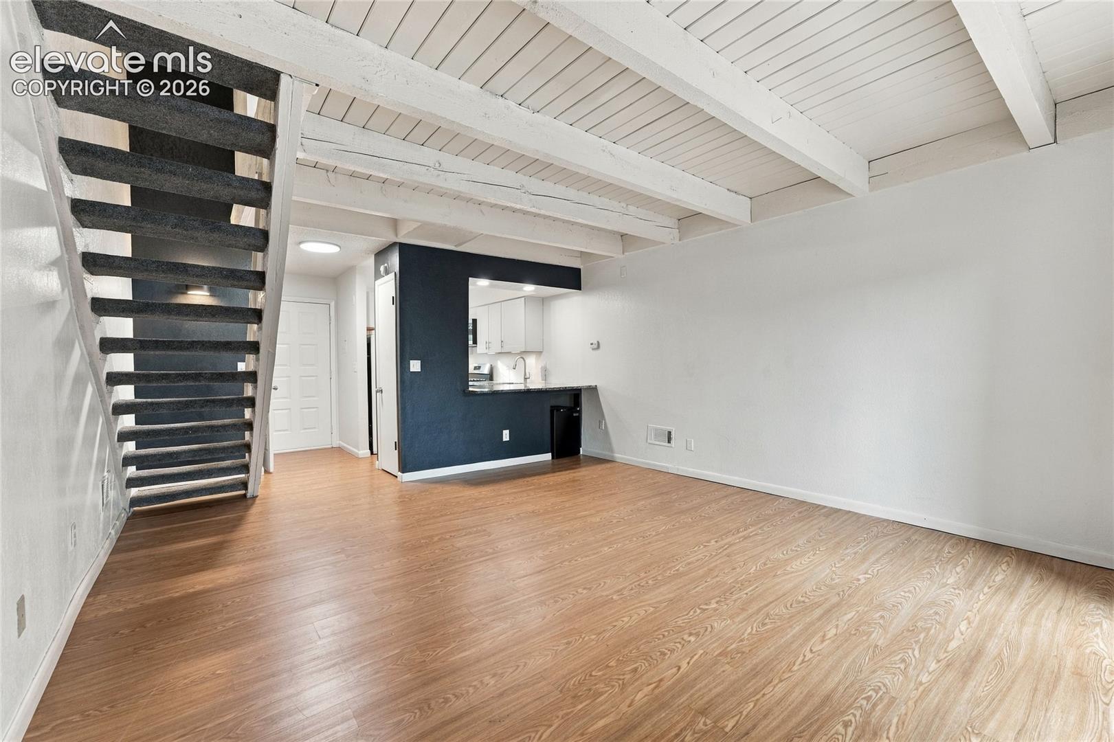 Open-concept interior featuring exposed white-painted timber ceiling beams, wood-finish flooring, a staircase with dark-toned treads, and a built-in kitchen pass-through with a dark-toned accent wall