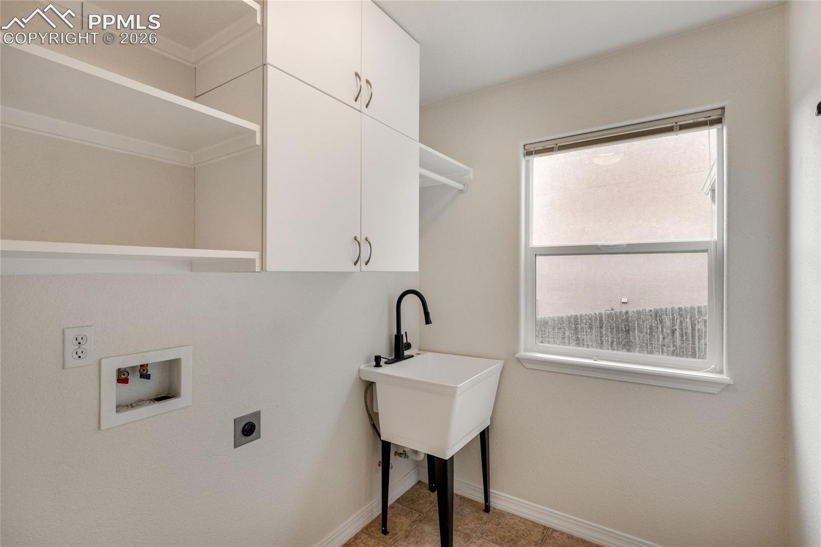 Dedicated laundry space featuring a utility sink with a black gooseneck faucet, white cabinetry with silver hardware, open shelving, and a window for natural light.
