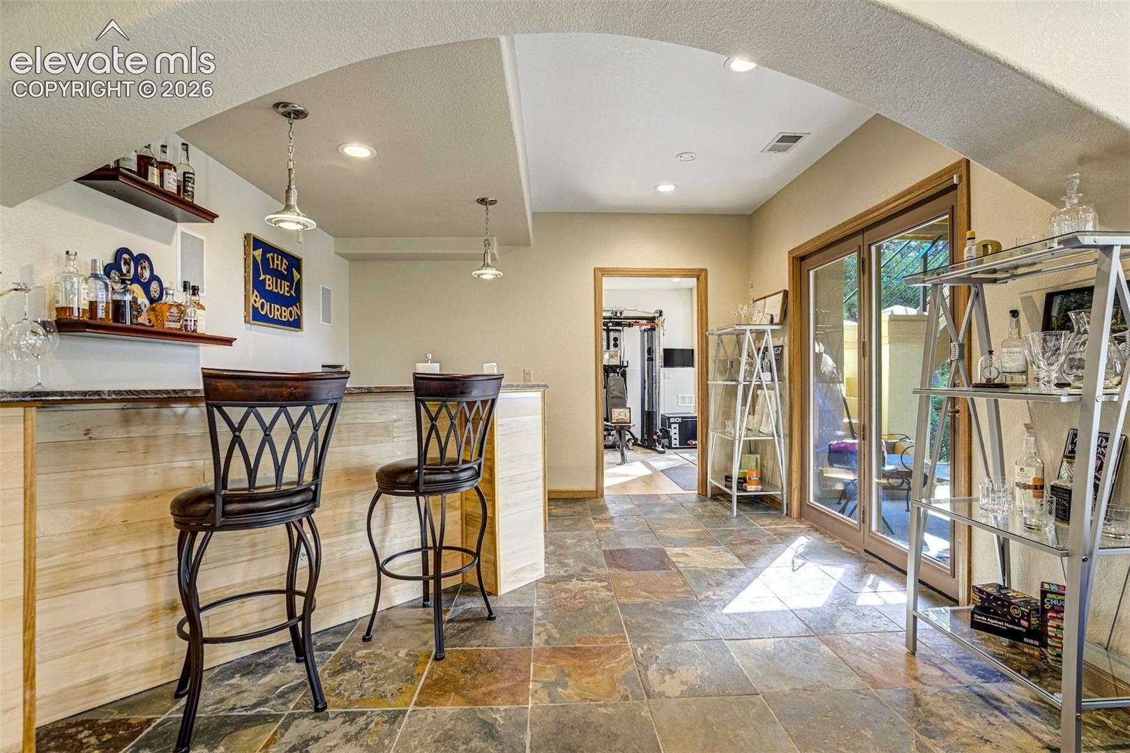 Built-in bar with wood-panel base and stone countertop, recessed lighting, pendant lighting fixtures, stone tile flooring, and glass-paneled double doors