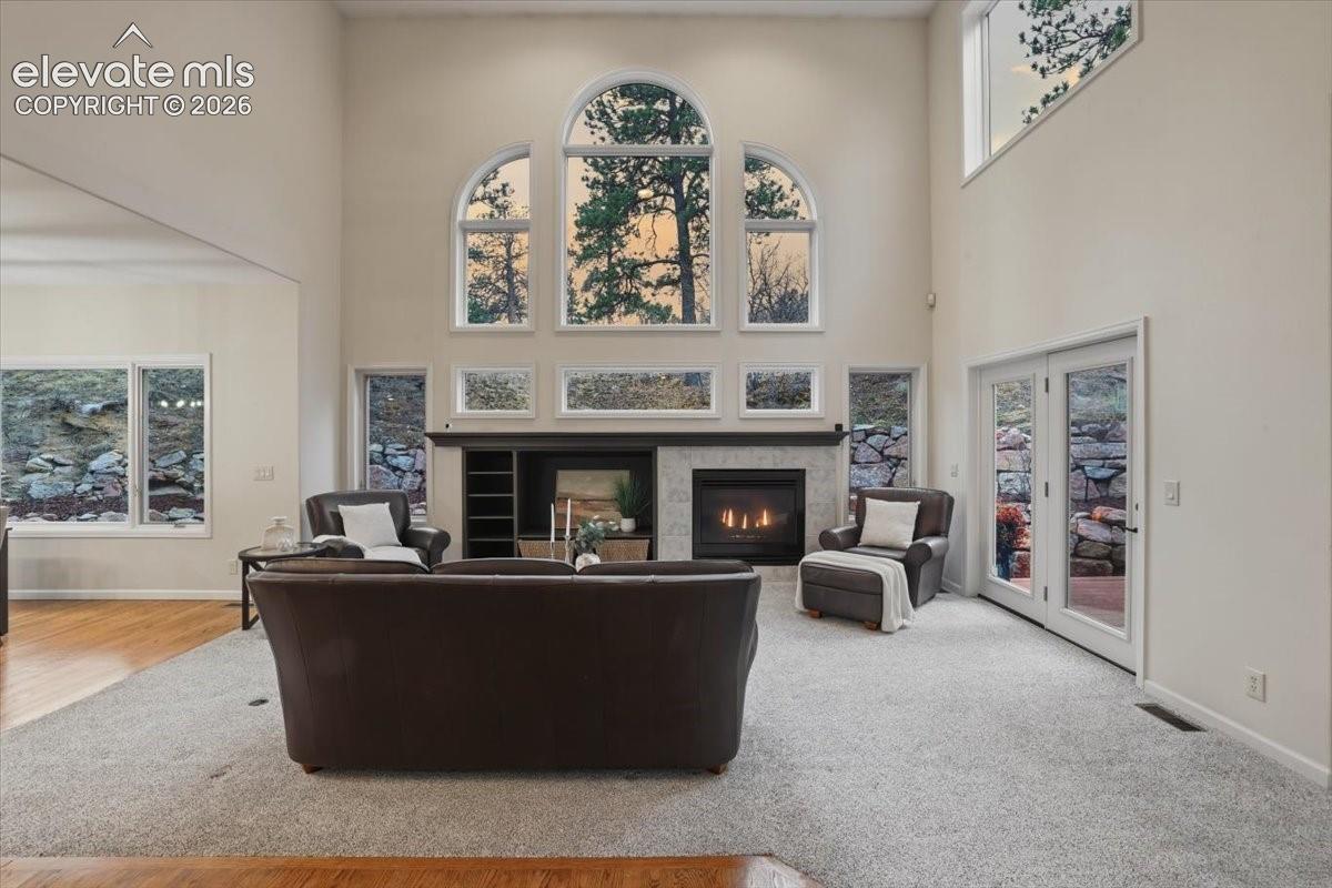 two story ceiling with arched windows, stone-clad fireplace with mantel, built-in shelving, light-colored carpeting, and French doors