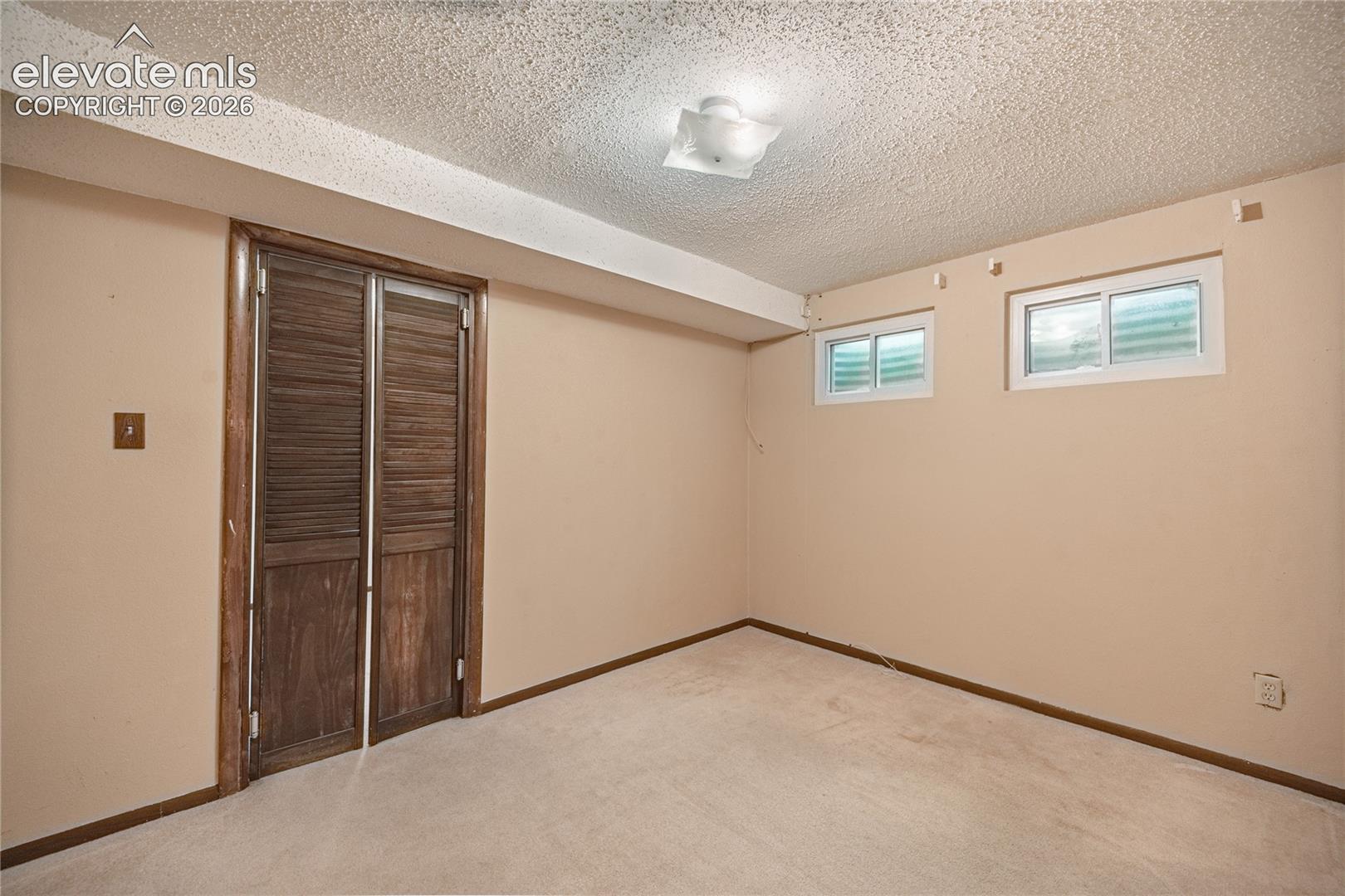 Interior space featuring dual-panel louvered closet doors, wall-to-wall carpeting, two horizontal egress windows, and a central ceiling-mounted light fixture