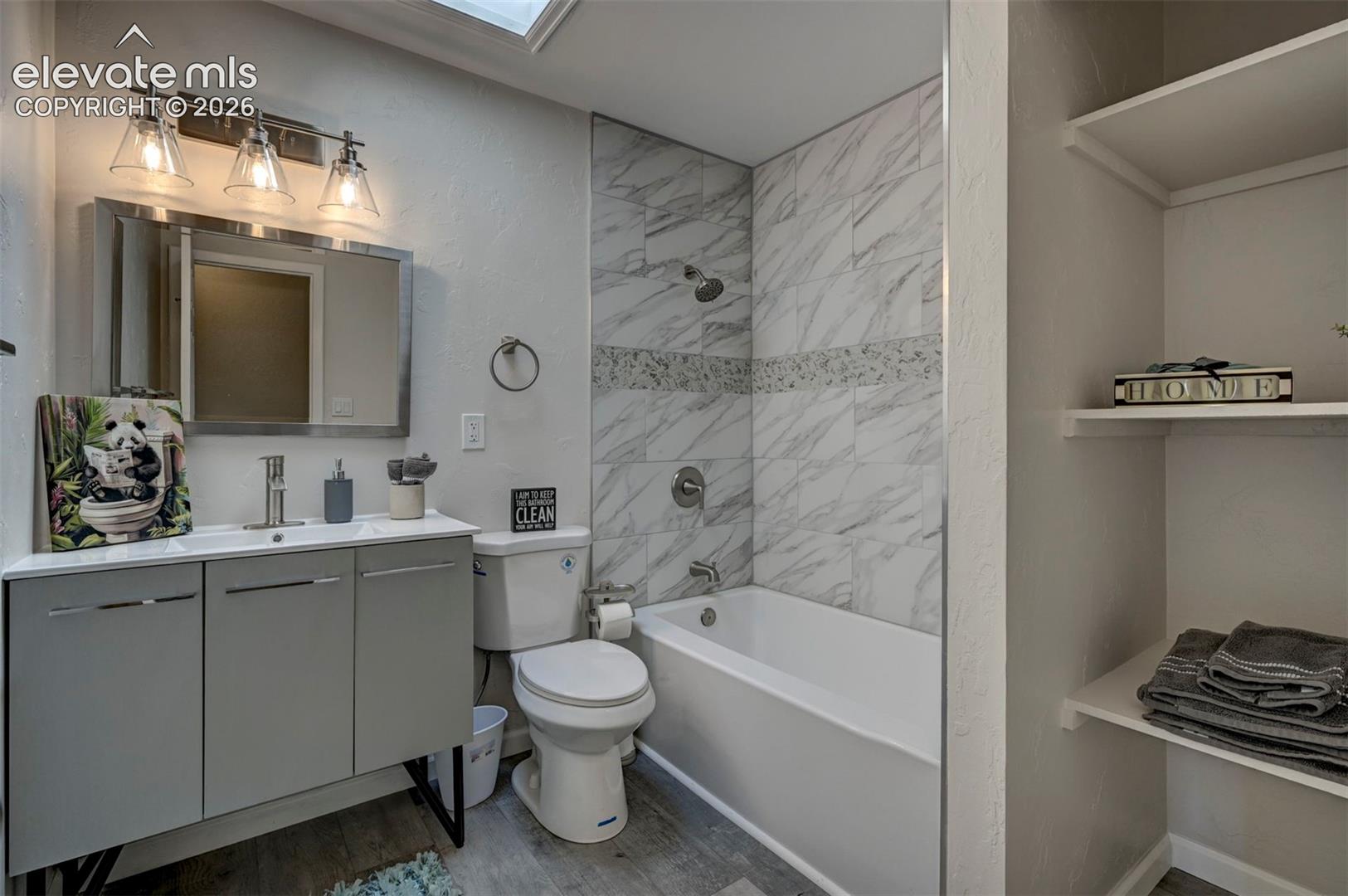Bathroom featuring a vanity with integrated sink, rectangular mirror with metallic frame, three-light fixture, ceiling skylight, and a tub enclosure with tile surround and decorative accent inlay
